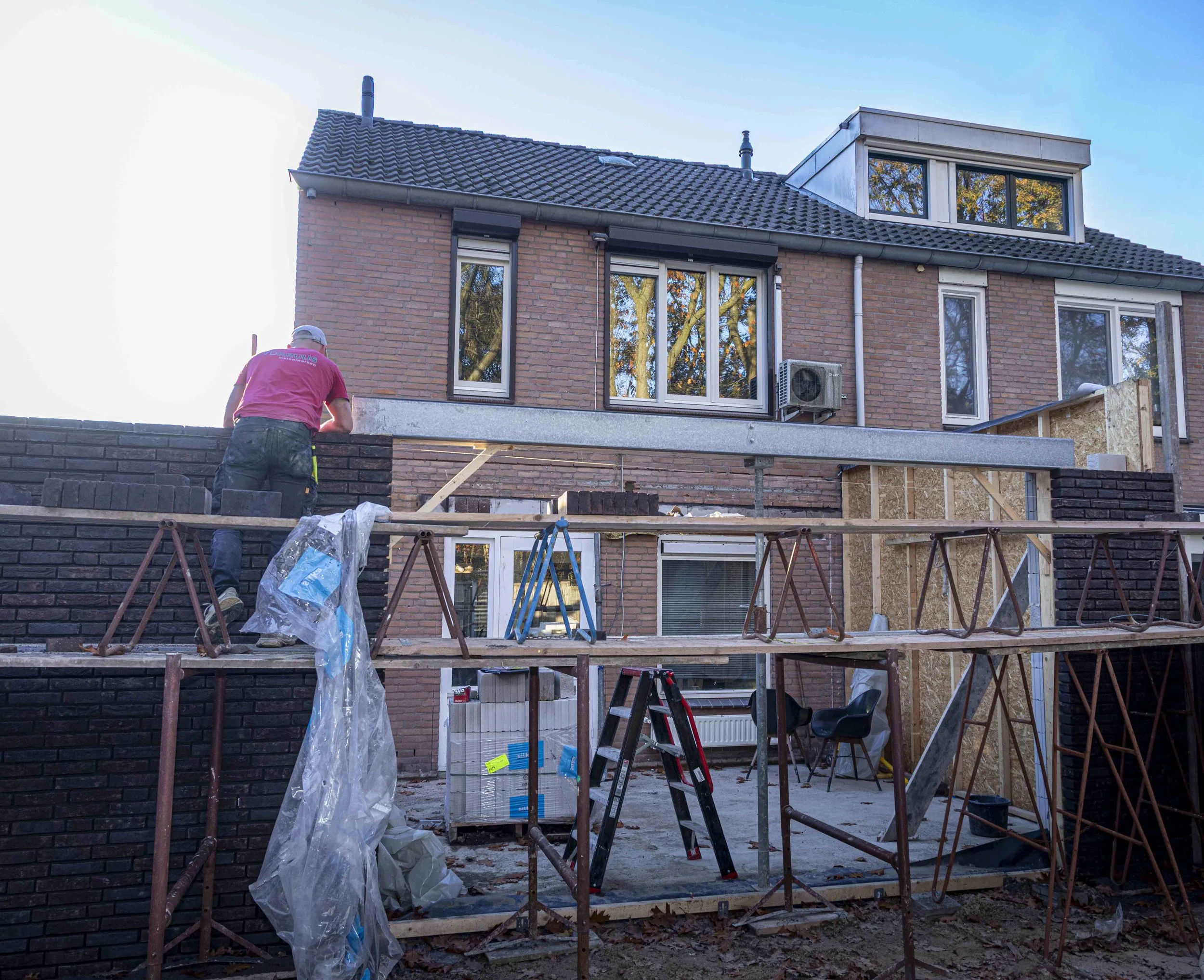 Een bouwvakker die een bakstenen muur metselt aan de buitenzijde van een meergezinswoning, met steigers en bouwgereedschap op de achtergrond.