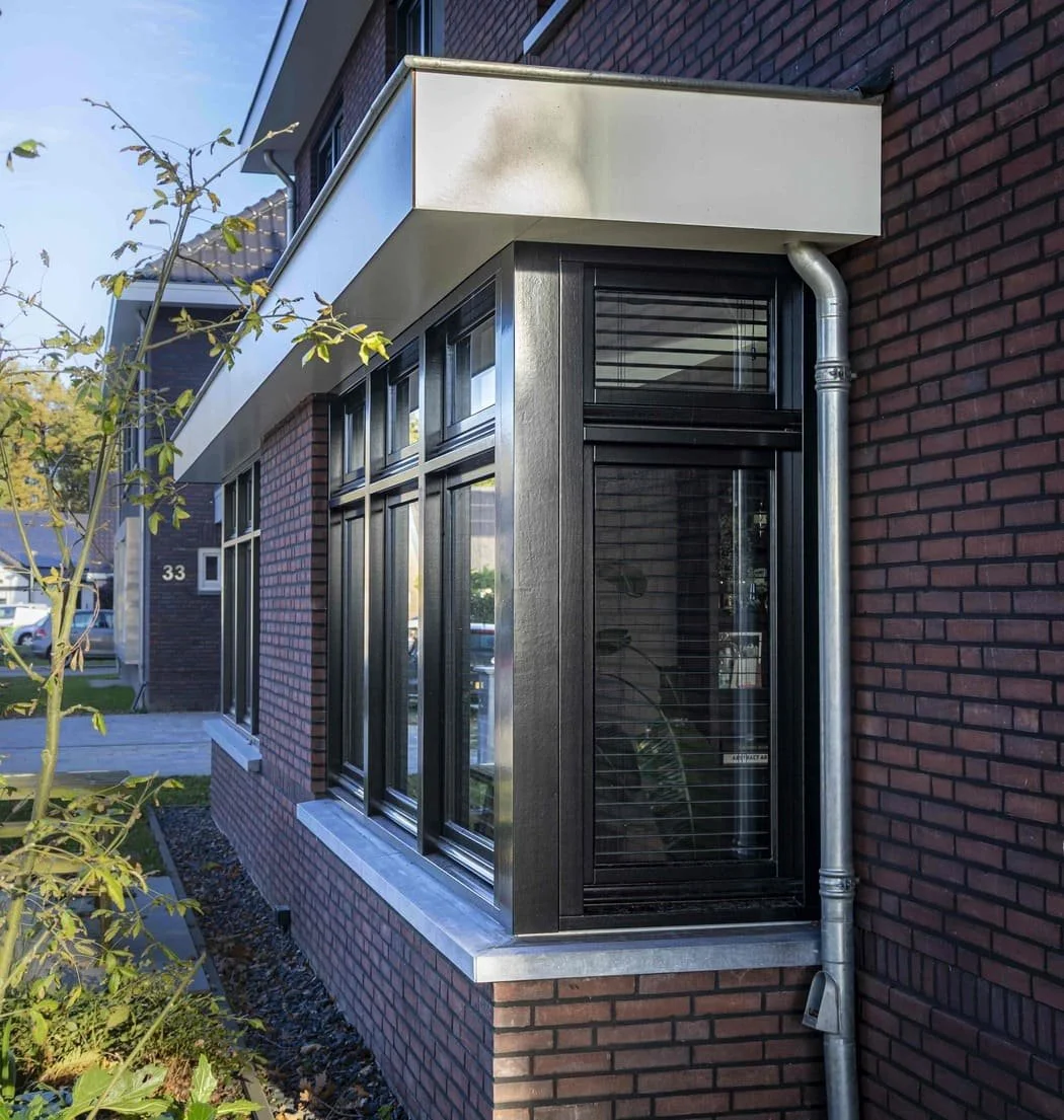 De foto toont een modern gebouw met een hoekige gevel, grote ramen met zwarte frames, een afwateringspijp, en een balkon met een witte rand op een bakstenen muur. Er is een boom en een tuin met grind ernaast.