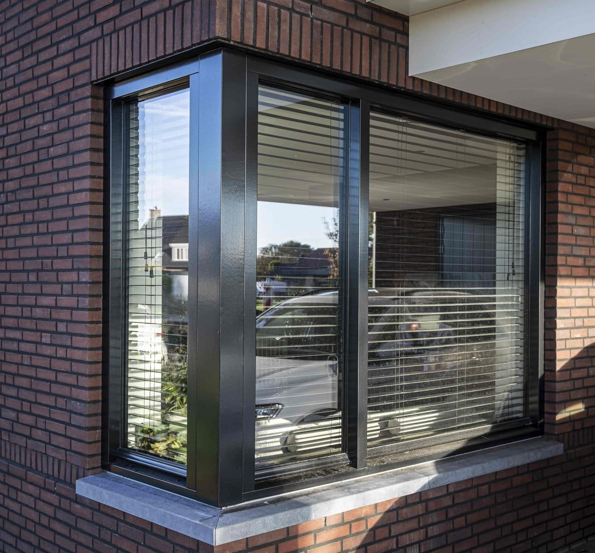 Een grote raampartij met gesloten horizontale jaloezieën, ingelijst door een zwarte metalen kader en geplaatst in een bakstenen muur, met reflecties van de omgeving.