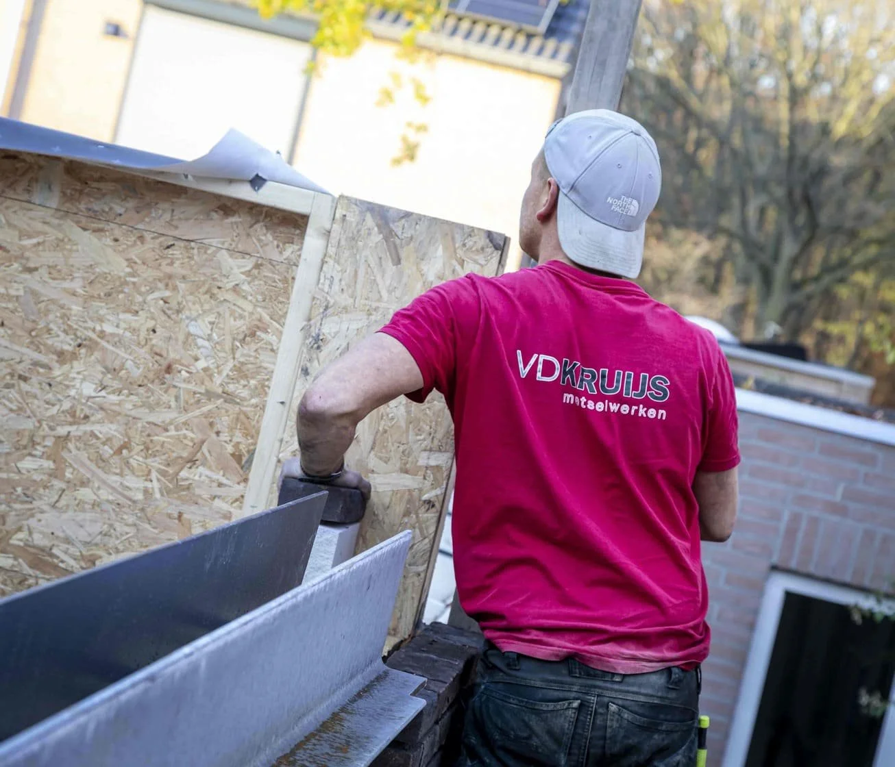 Aannemer in roze T-shirt en witte pet die werkt aan bouwproject, met hout en bouwmaterialen op de achtergrond.