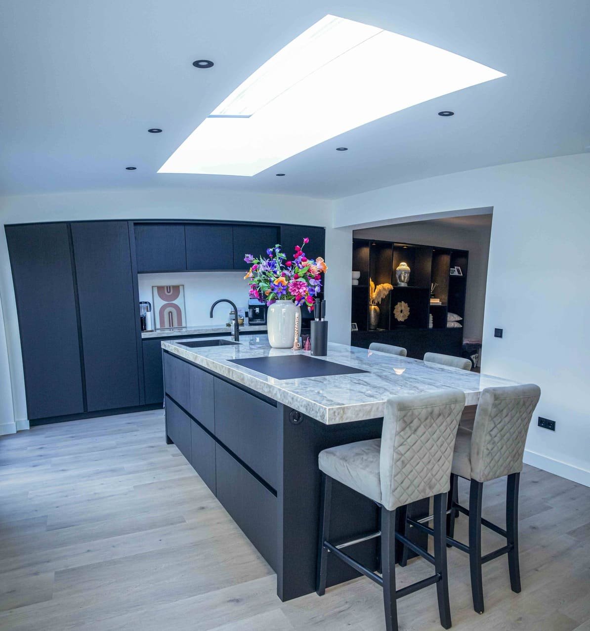 Moderne keuken met een eiland, marmeren werkblad en zwarte kasten. Een grote vaas met kleurrijke bloemen staat op het eiland, en er zijn beige quastenstoelen. Het plafond heeft een grote lichtkoepel.