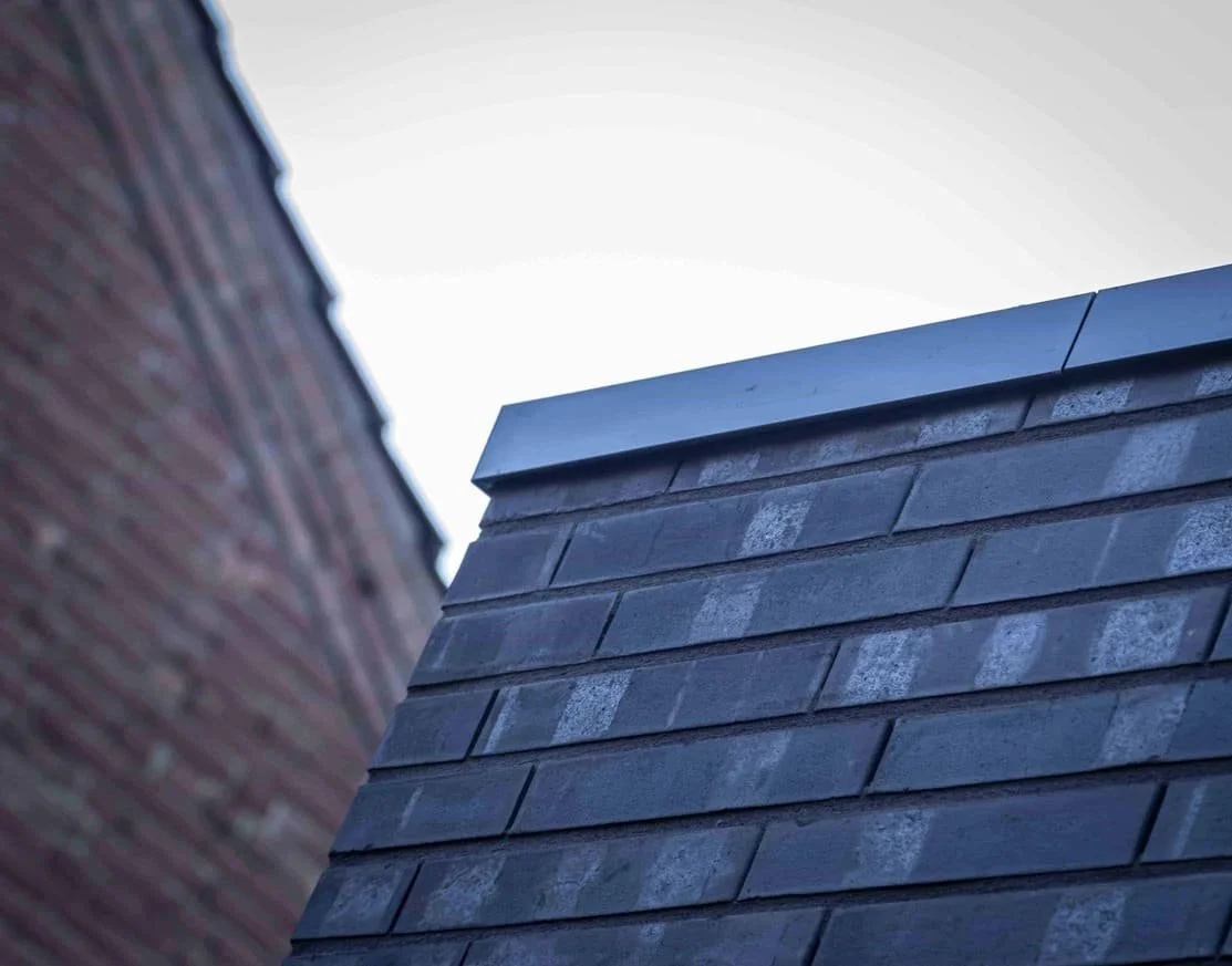Close-up van een dak met dakpannen, gezien vanaf beneden met een gedeelte van een andere muur op de linkerzijde en de lucht op de achtergrond.