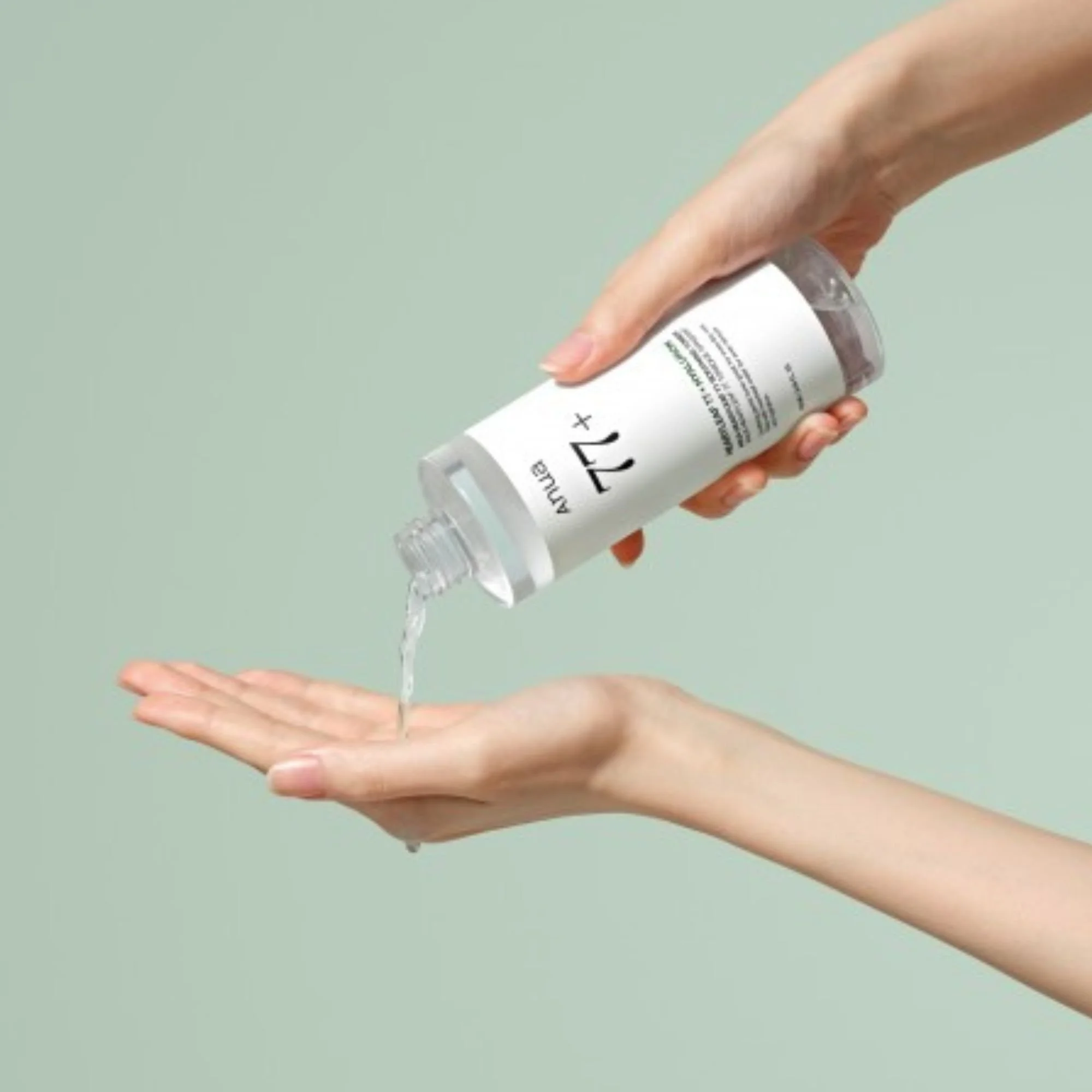 Mains versant le Anua Heartleaf 77% + Hyaluron Soothing Toner dans la paume. Application du tonique K-Beauty hydratant sur fond vert menthe.
