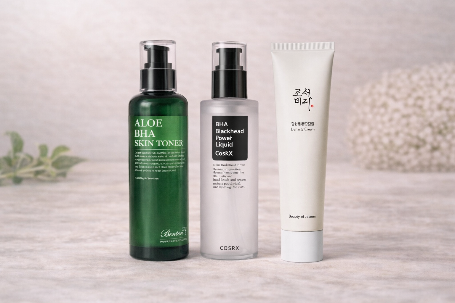 Trois produits de soin de la peau disposés sur une surface en bois clair avec un fond flou. Le premier est un flacon vert intitulé "ALOE BHA SKIN TONER", le second un flacon blanc avec un étiquette noir intitulé "BHA Blackhead Power Liquid CosRX", et le troisième un tube blanc nommé "Dynasty Cream".