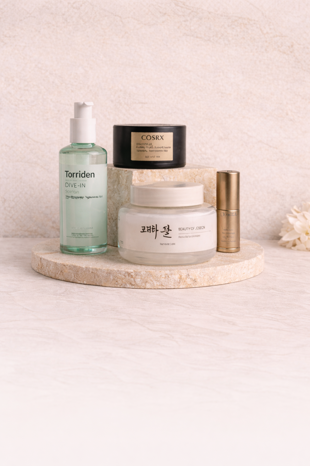 Collection de produits de soin sur une plateforme en pierre, comprenant une bouteille d'eau micellaire, une crème en pot, un pot noir et un petit tube doré, avec un fond neutre et floral.