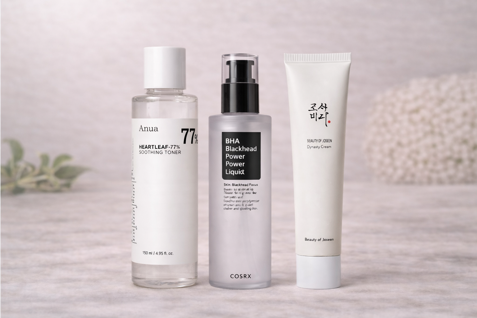 Trois produits cosmétiques alignés : un toner transparent avec un capuchon blanc, un liquide noir dans un flacon gris avec un pulvérisateur noir et un tube blanc avec des écritures en coréen et en anglais.