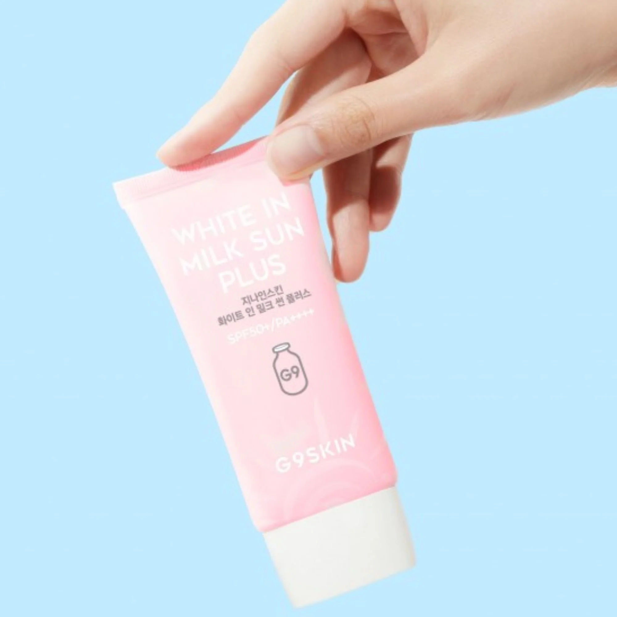 Main tenant le tube de G9SKIN White In Milk Sun Plus SPF50+ PA++++ sur fond bleu. Crème solaire K-Beauty rose pâle.