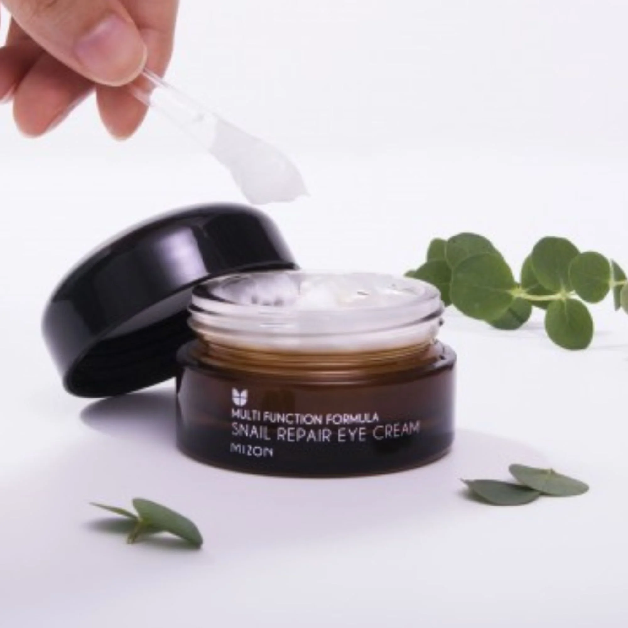 Texture du Mizon Snail Repair Eye Cream. Crème pour les yeux K-Beauty à la bave d'escargot prélevée à la spatule dans le pot ambré, avec feuilles d'eucalyptus.