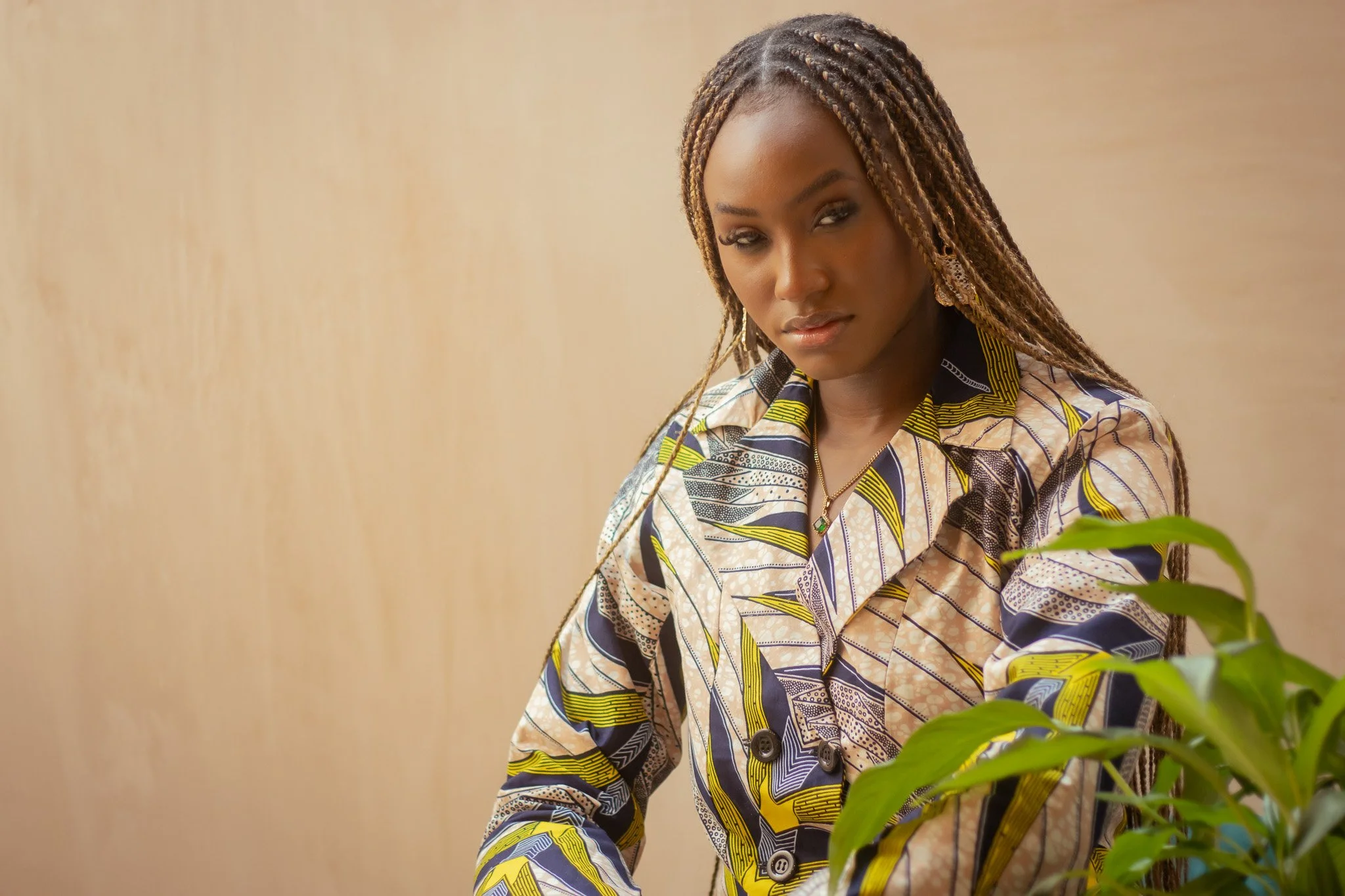 Cinematic mid-shot of a woman with braids wearing an African print suit, using a neutral studio backdrop, organic greenery and patterned styling to create depth and visual harmony — signature portrait cinematography by ABHIOLAR.ART.