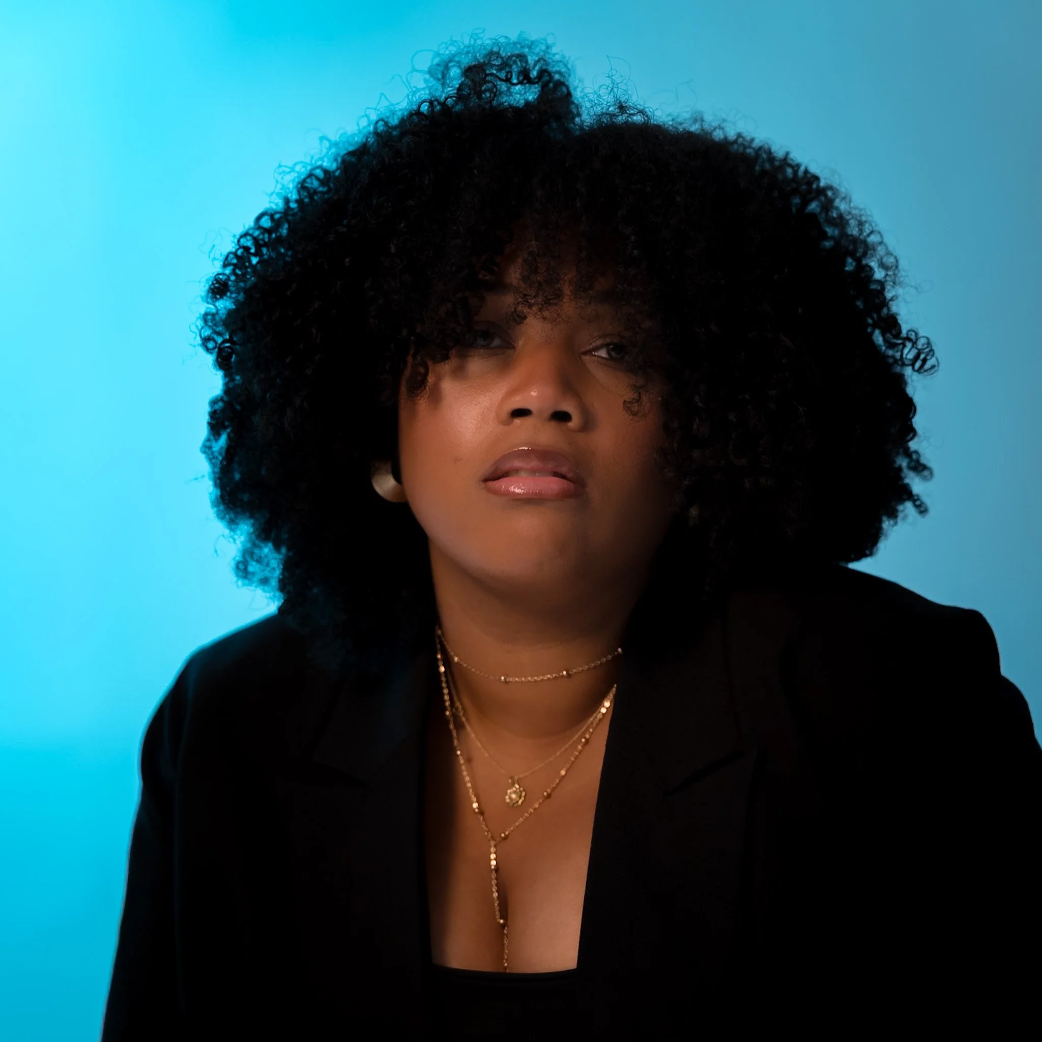Cinematic portrait of a woman with curly black hair wearing a black blazer and layered gold necklaces and earrings, set against a blue backdrop — editorial visual storytelling by ABHIOLAR.ART.
