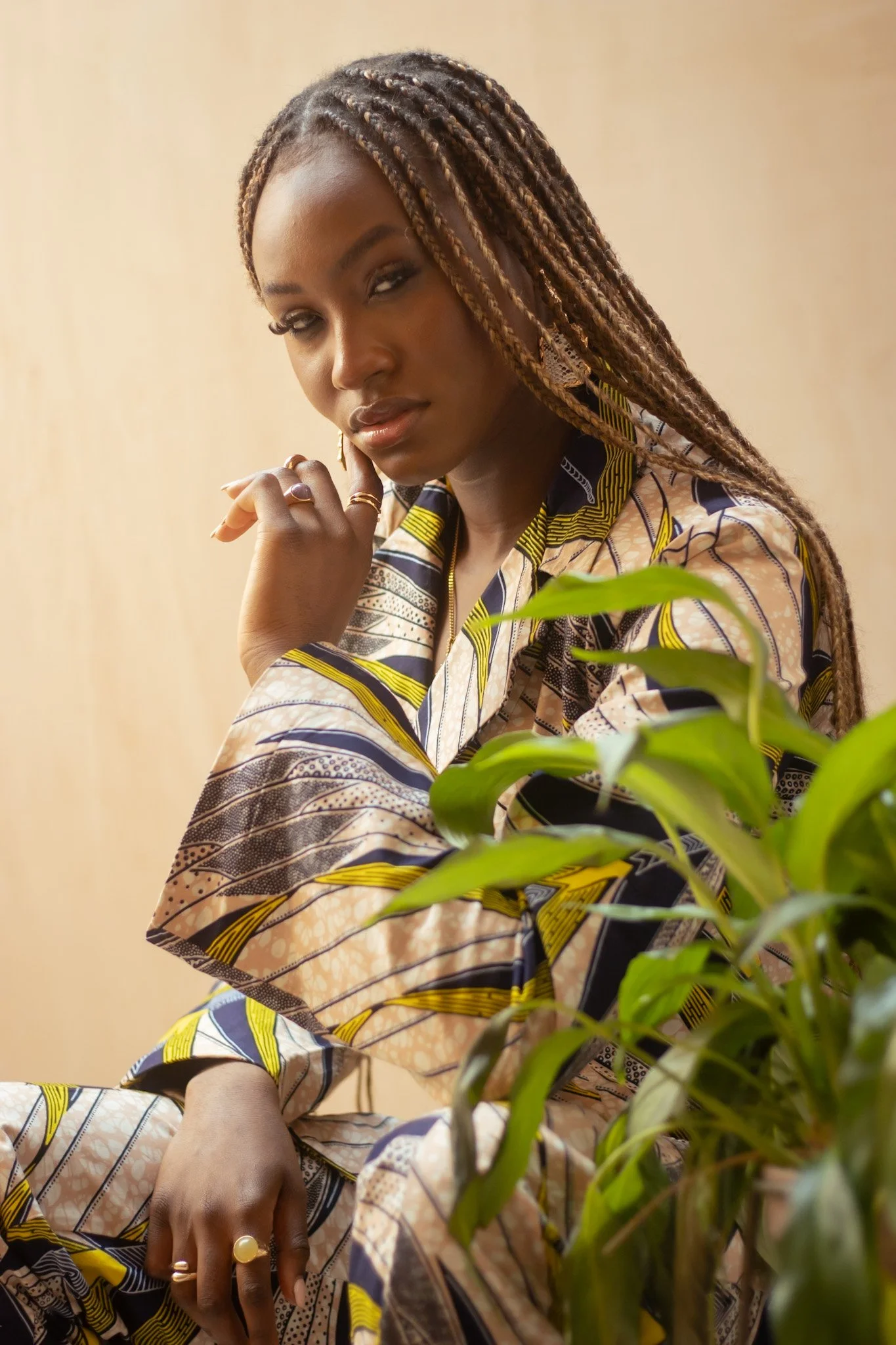 Cinematic close-up of a woman with braids, wearing an African print suit, using soft directional light, a thoughtful hand gesture and organic greenery to shape depth and tone — signature portrait cinematography by ABHIOLAR.ART.