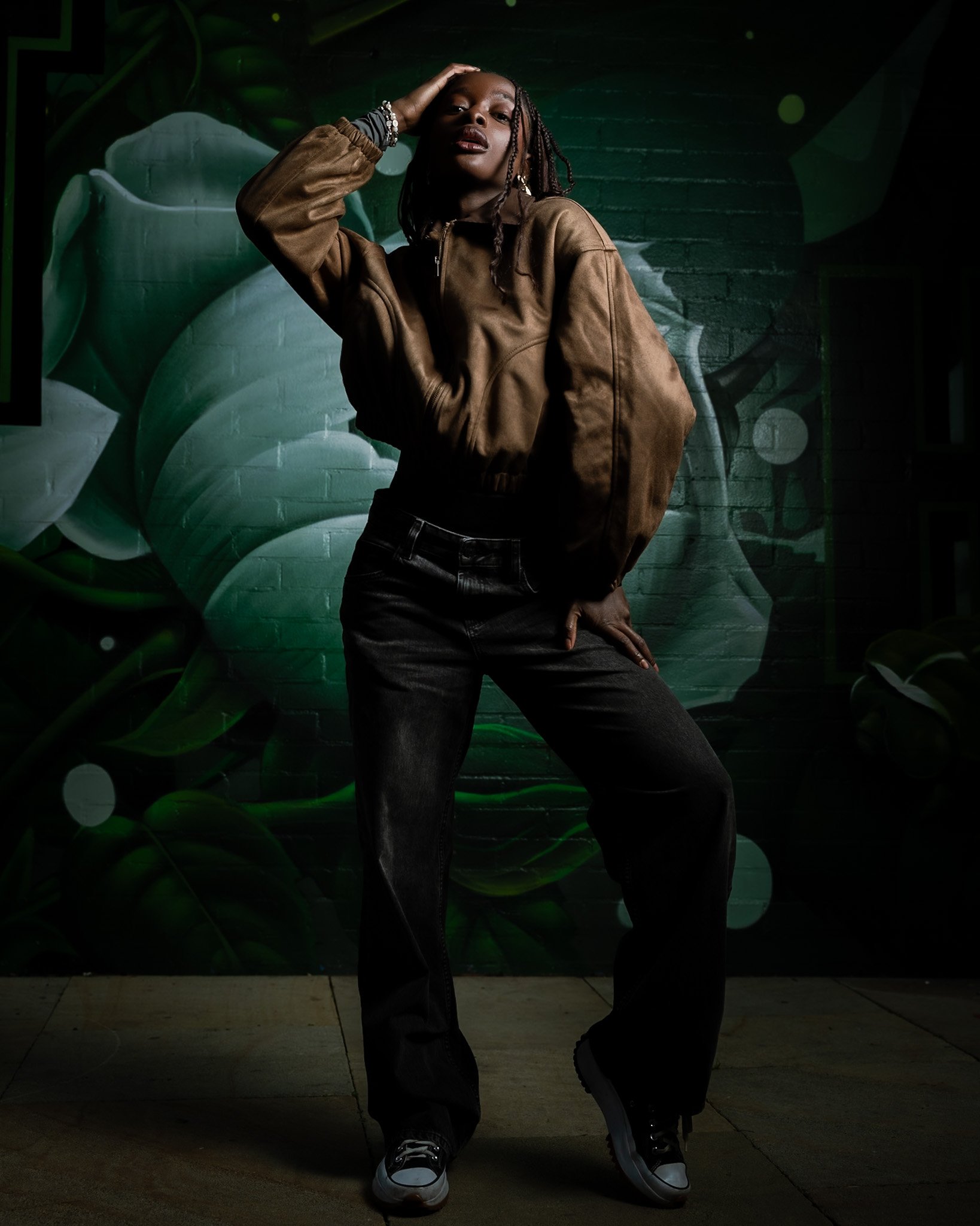 Cinematic full-body portrait of a woman with braids, using natural light, rich green mural textures and strong posing to create depth and presence — signature cinematography by ABHIOLAR.ART.