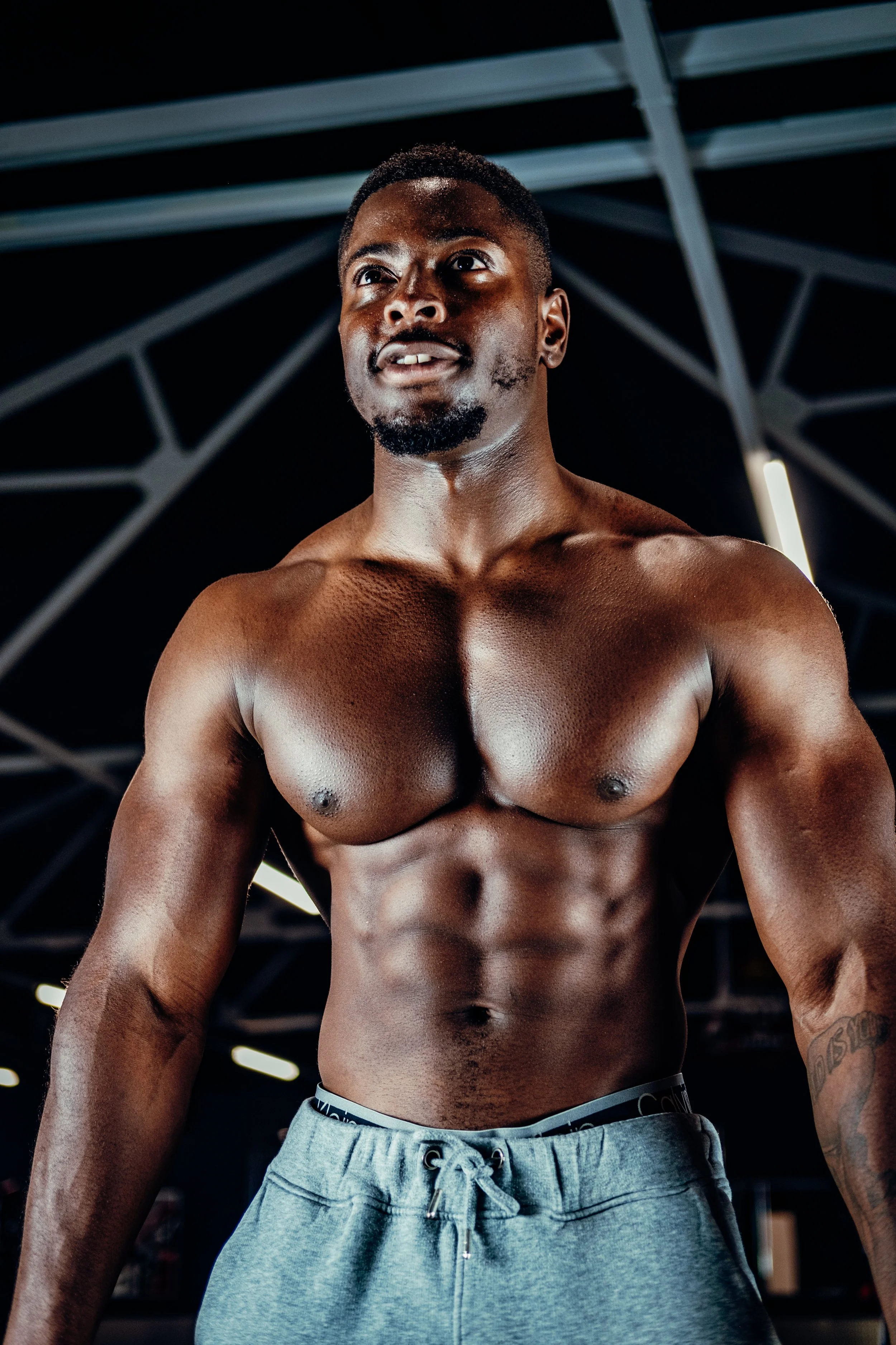 A muscular man standing shirtless in a gym with a dark ceiling and overhead lighting.