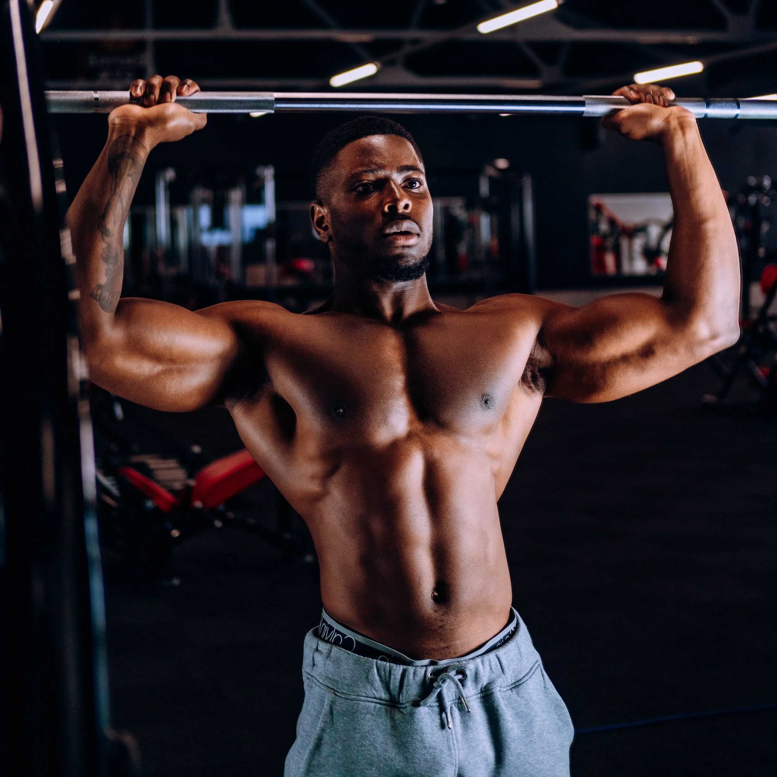 A muscular man with dark skin lifting a barbell in a gym, shirtless and wearing grey sweatpants.