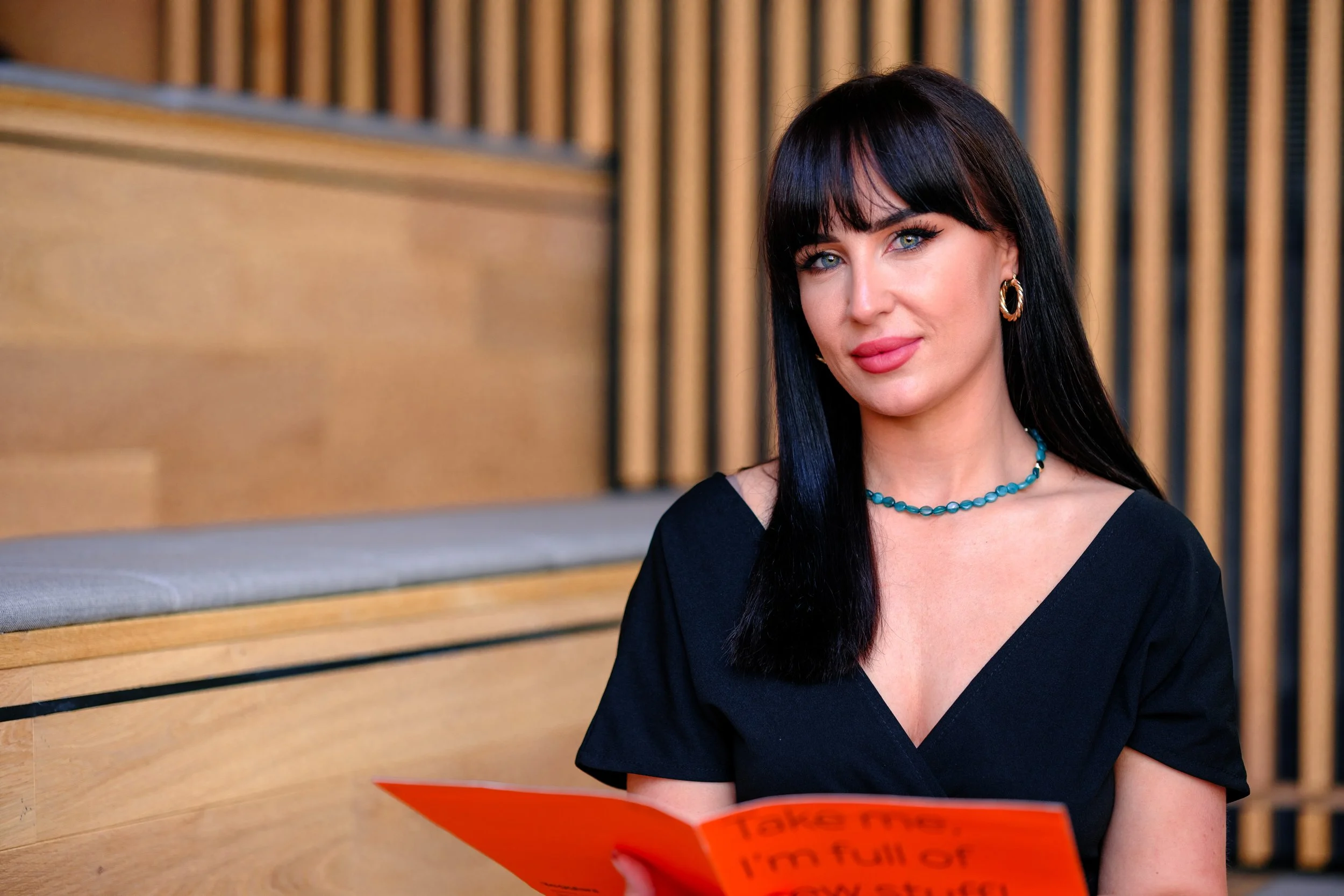 A woman with long black hair and blue eyes, wearing a black top, gold hoop earrings, and a blue beaded necklace, is sitting and holding an orange booklet with red text, in a wooden interior setting.