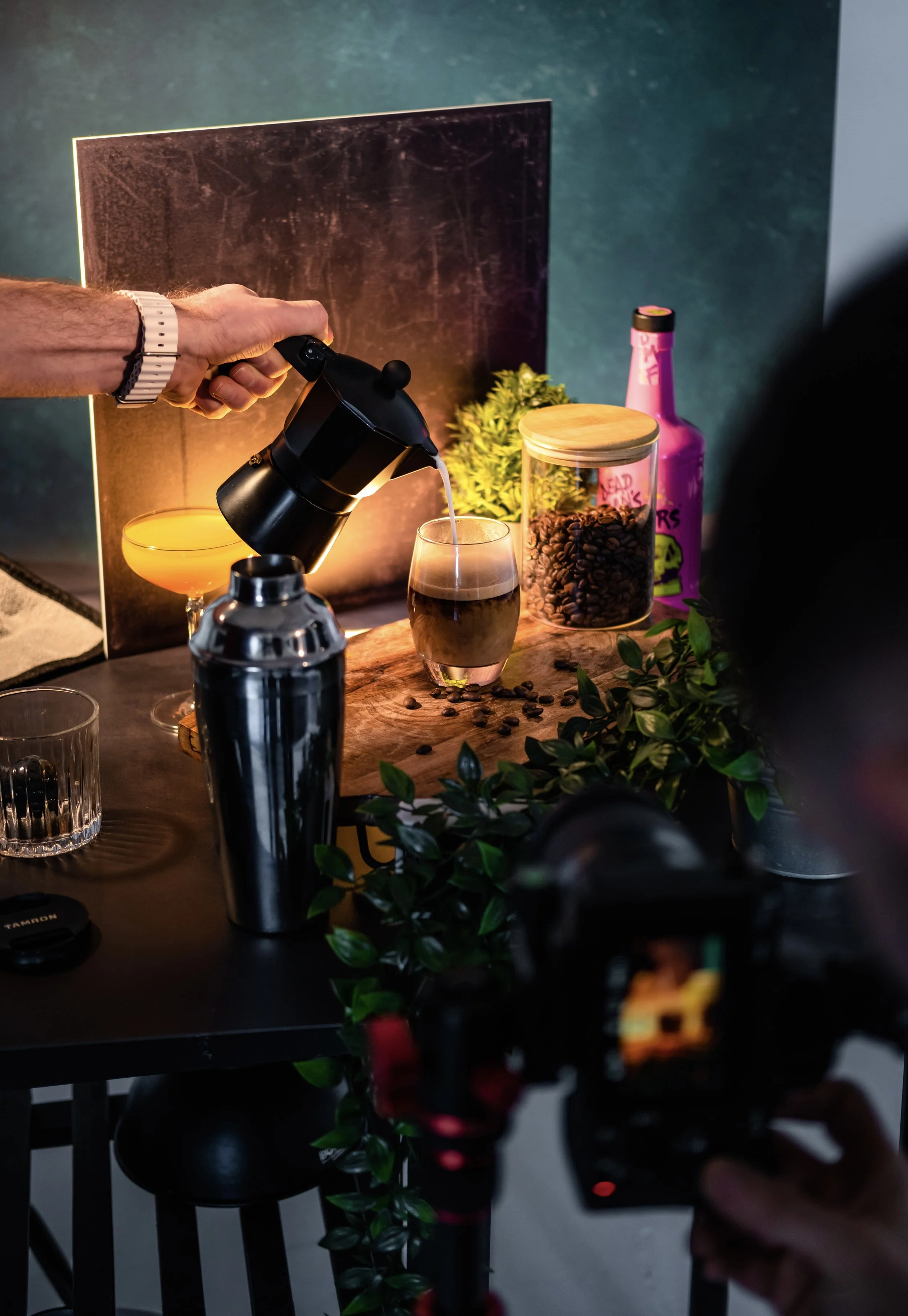 A person is pouring coffee from a French press into a glass. The scene is set on a table with various items, including a cocktail glass, a canister of coffee beans, a plant, a pink bottle, and a black camera in the foreground capturing the scene.