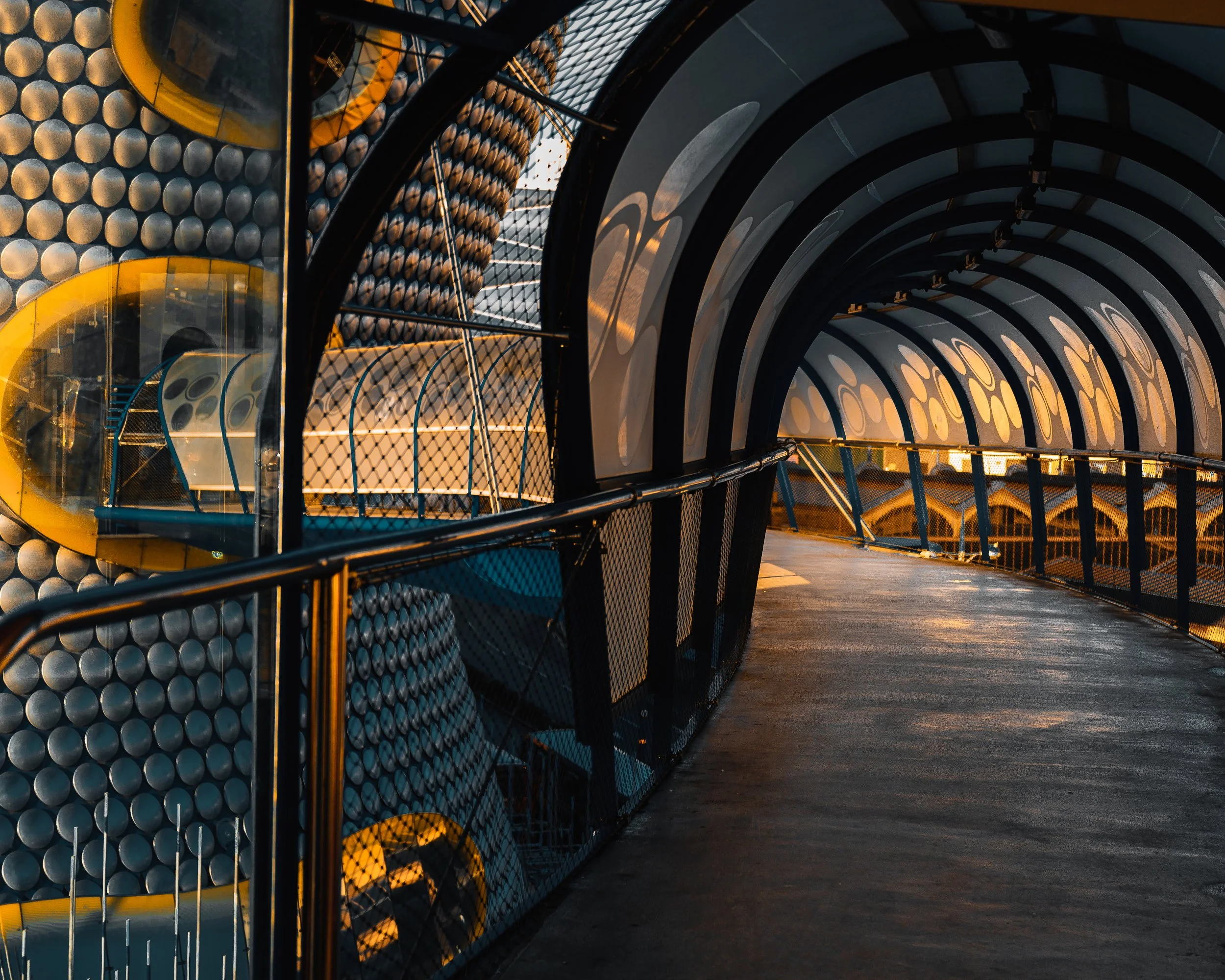 Futuristic outdoor walkway with curved glass and metal structure, illuminated by warm sunlight, featuring creative architectural design with circular and cylindrical elements.