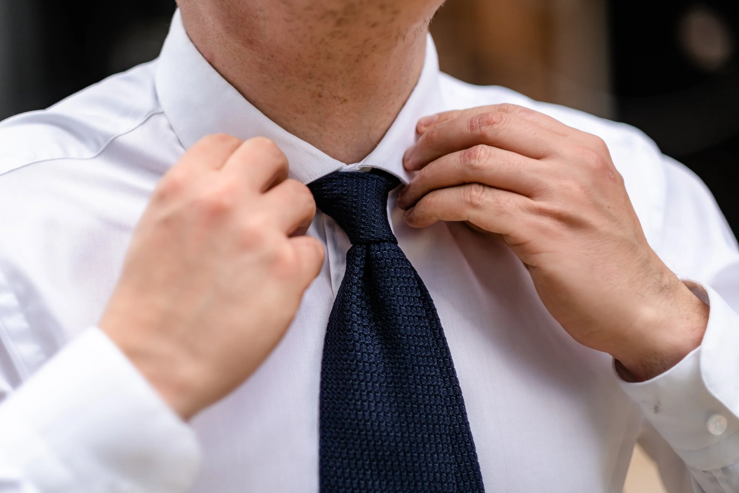 A man in a white shirt adjusting a navy blue tie.