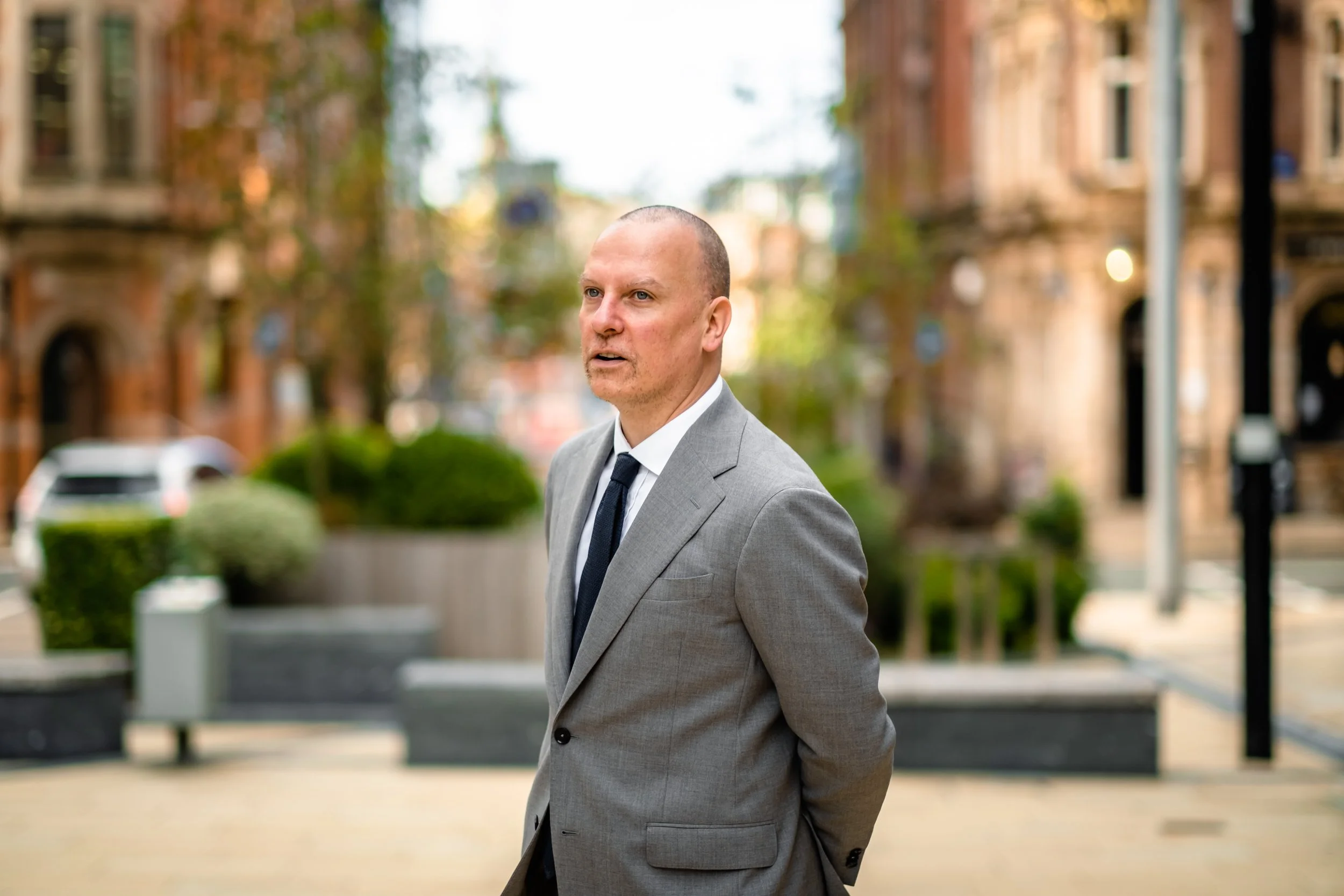 A man in a gray suit with a white shirt and black tie, standing outdoors on a city street with buildings, trees, and parked cars in the background.