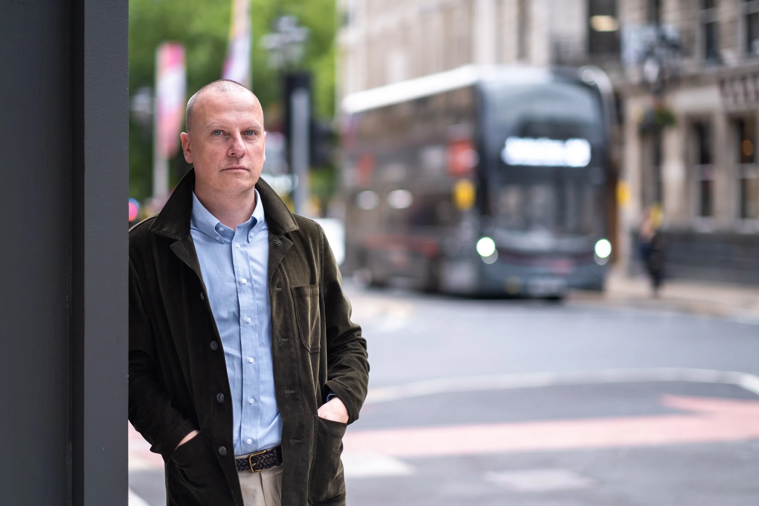 A man in a brown coat and light blue shirt standing on a city street, leaning against a wall, with a bus in the background.