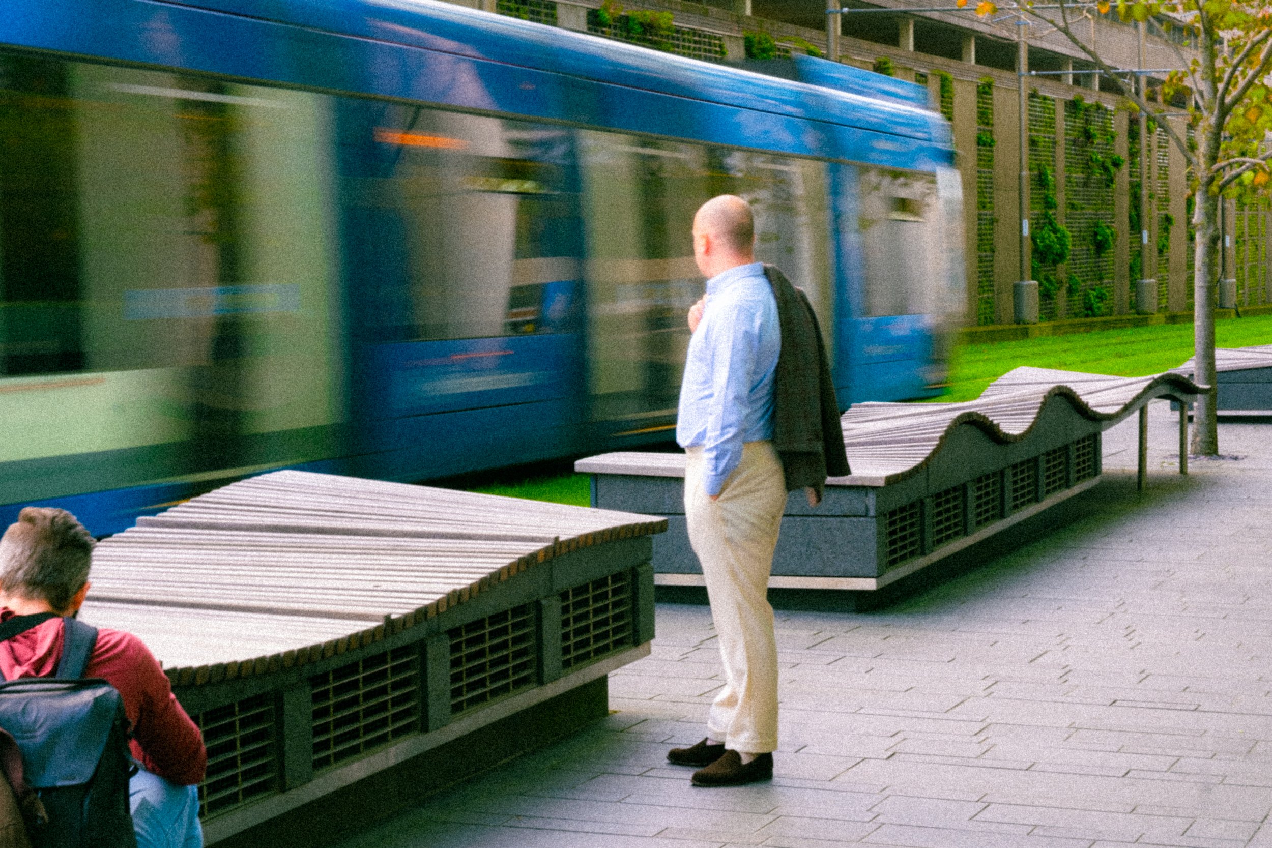 A man standing on a sidewalk in front of a moving blue train, with another person sitting nearby on a bench.