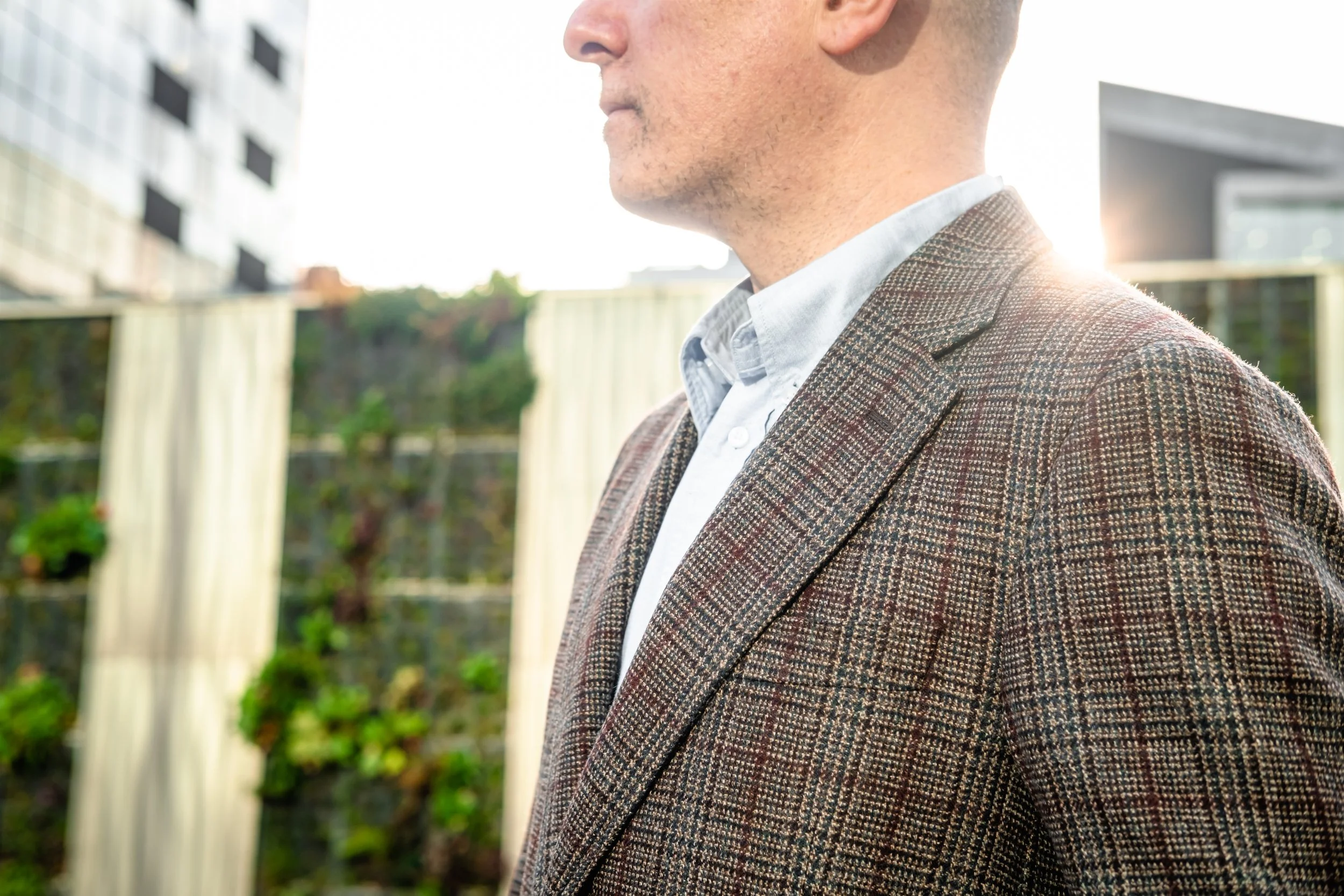 Close-up side view of a man wearing a brown plaid blazer and a white shirt, outdoors with buildings and a garden in the background.