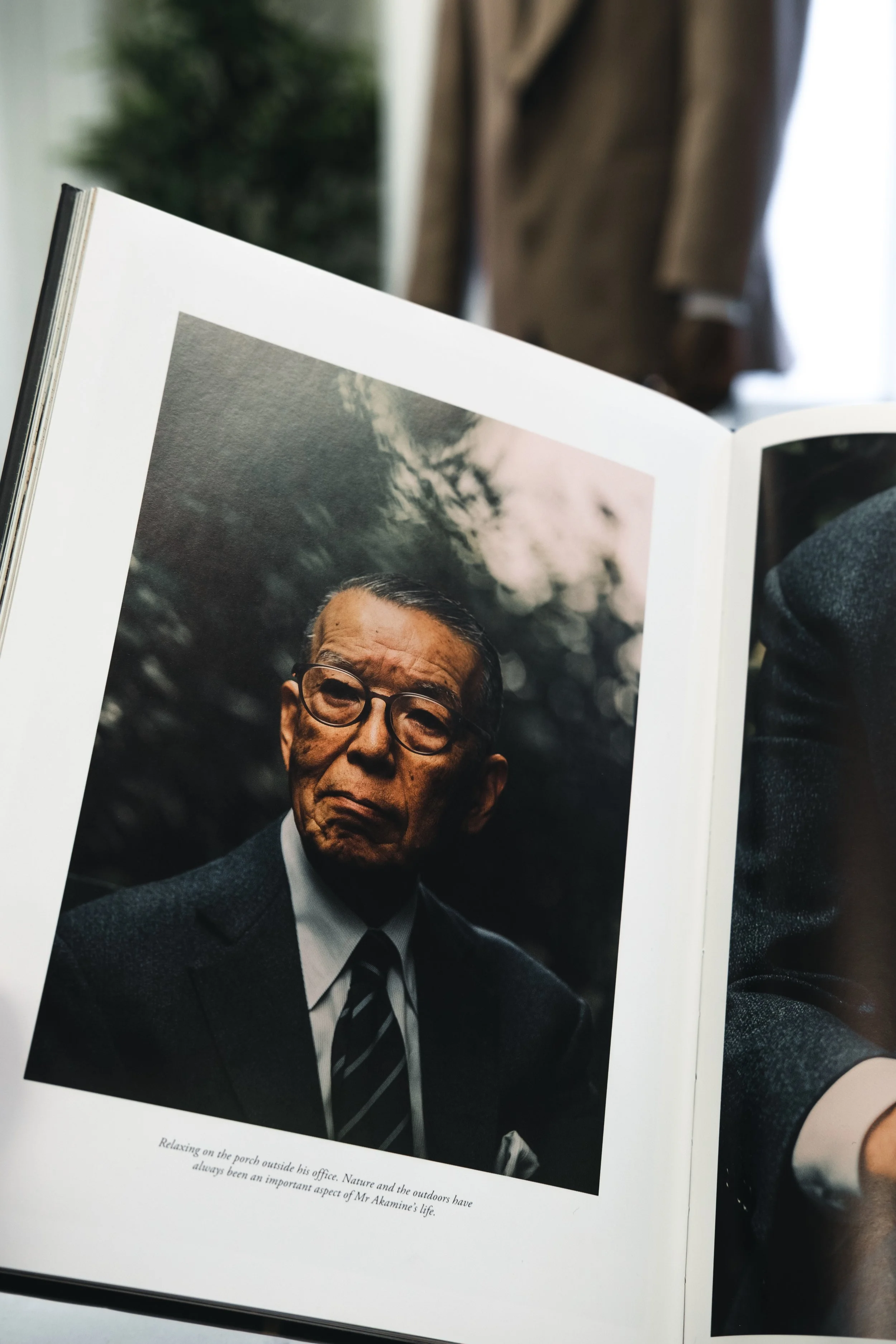 A man in glasses wearing a suit and tie, looking serious, outdoors with trees and clouds in the background.