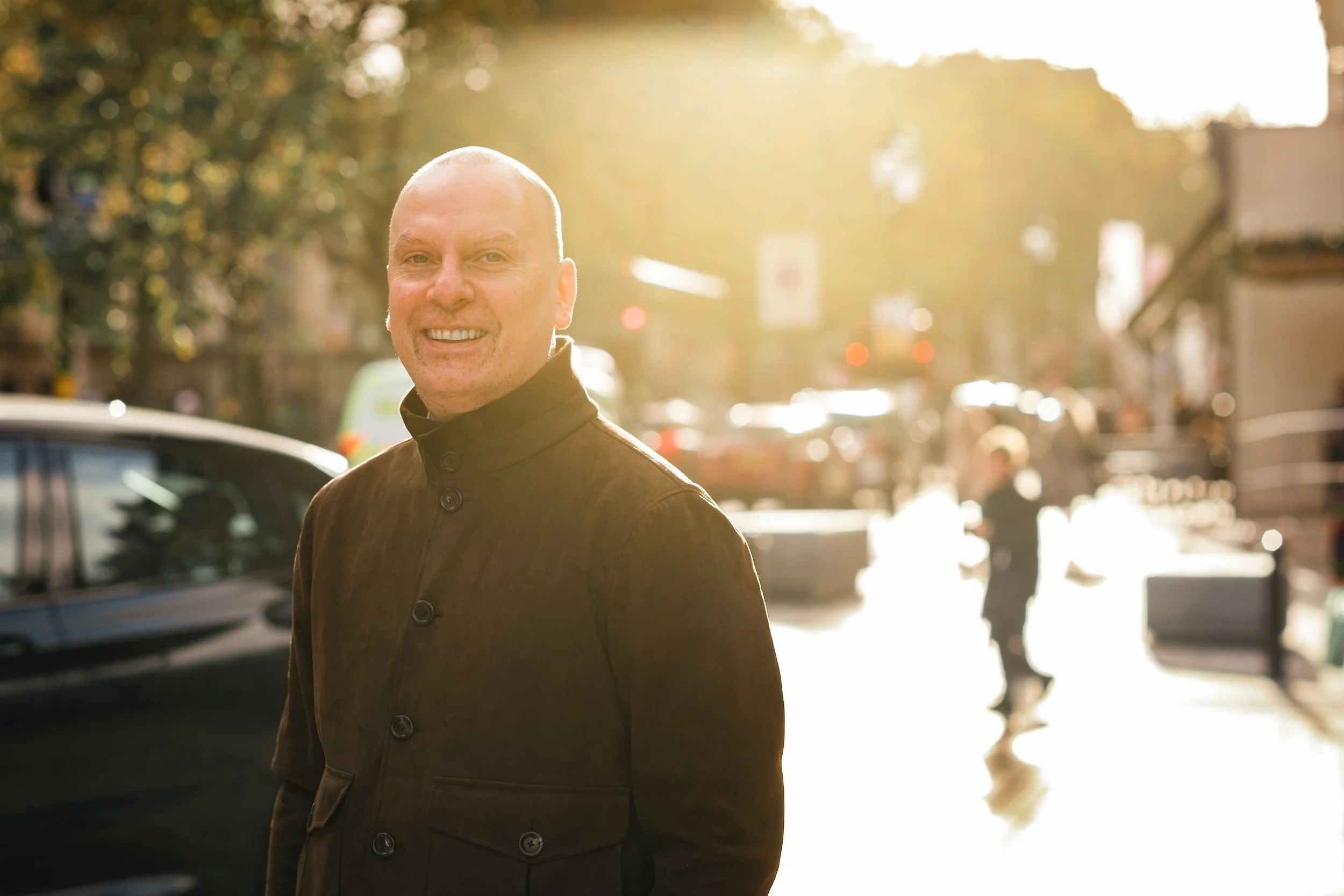 A smiling man with a shaved head and beard, wearing a dark coat, standing outdoors in a city street during sunset.