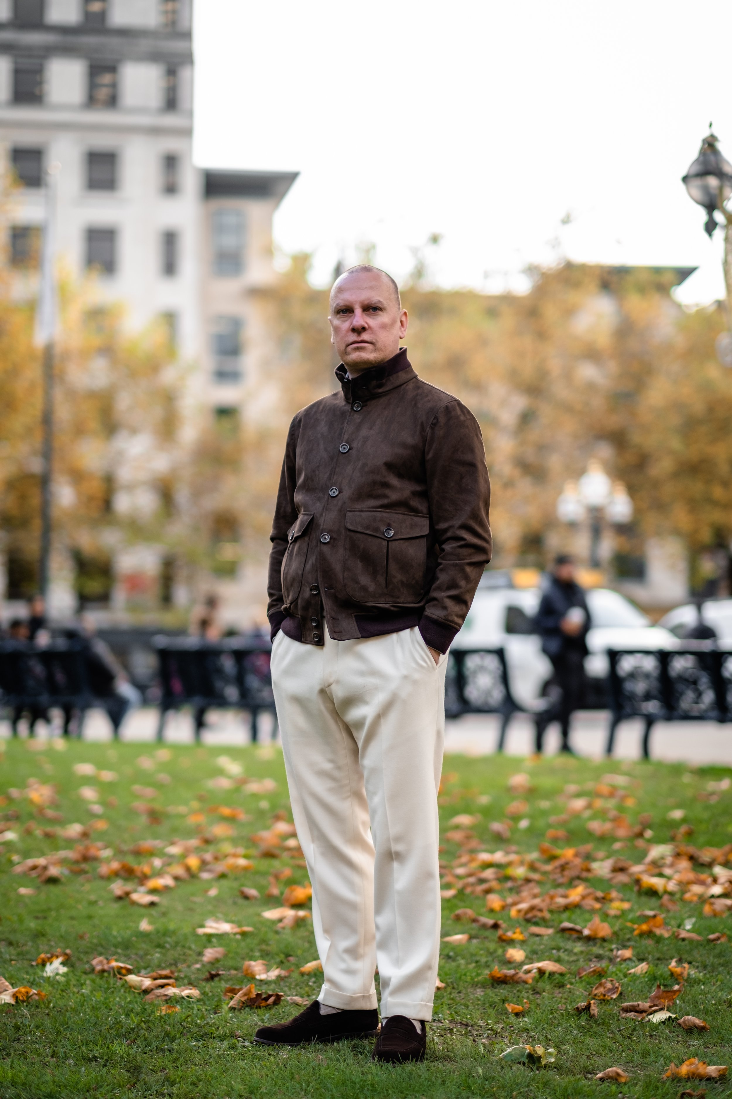 A man standing in a park with autumn leaves on the ground, wearing a brown jacket and white pants, with buildings and other people in the background.