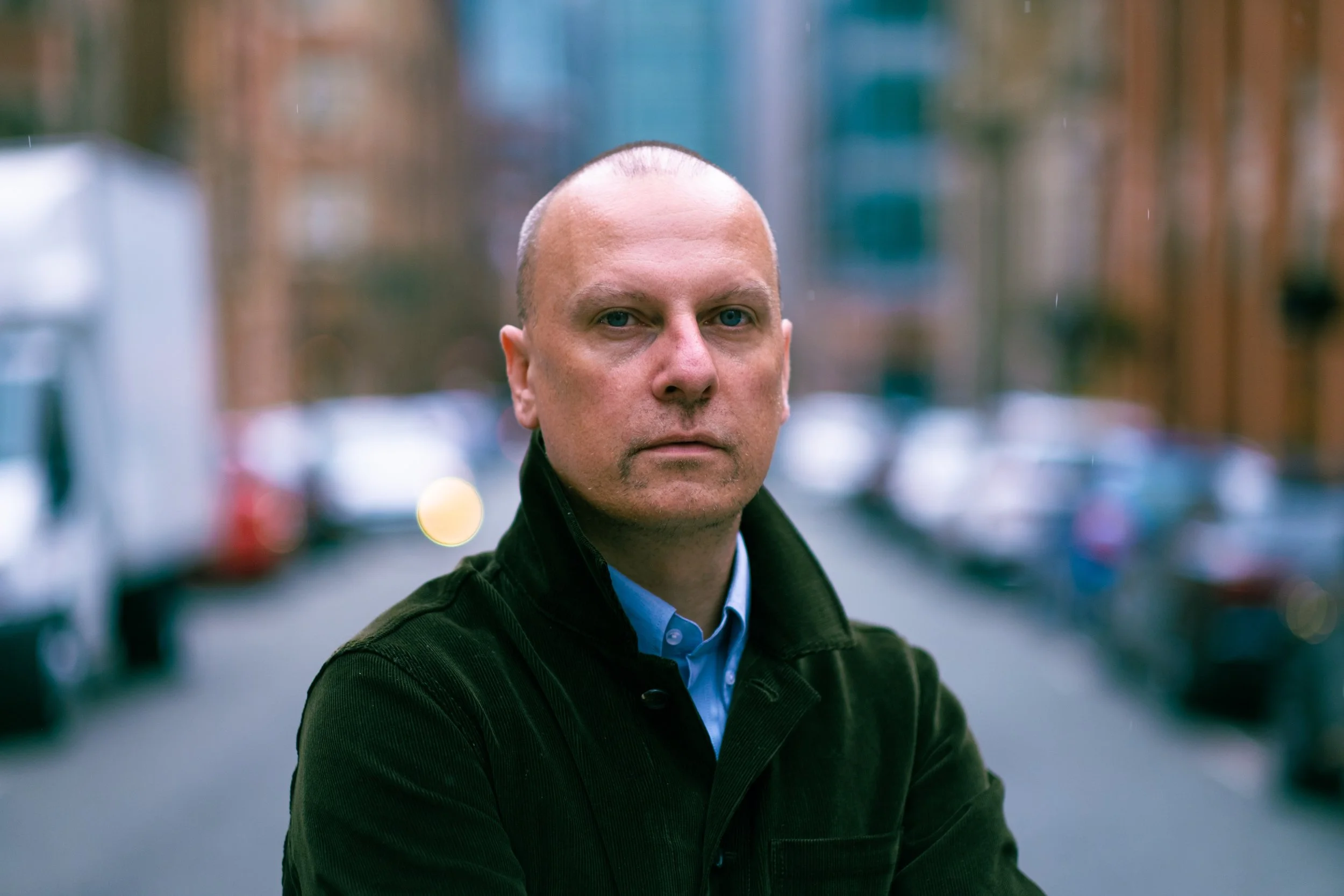 A bald man with light skin and blue eyes wearing a black jacket and blue shirt, standing outdoors on a city street with blurred cars and buildings in the background.
