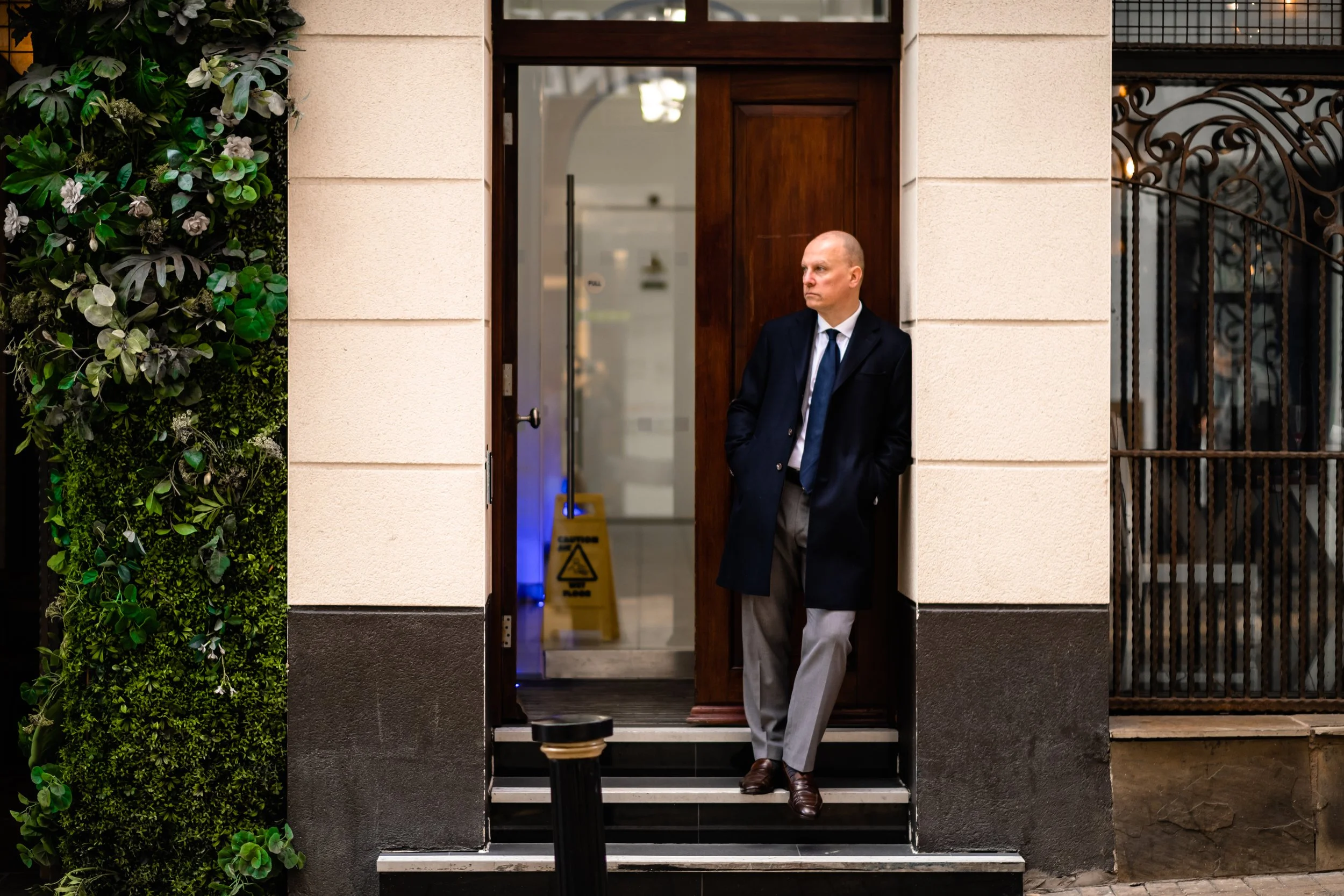 A man in a business suit with a dark coat and grey trousers standing on the front steps of a building with his hands in his pockets and looking to the side. The building has beige walls, a dark wooden door, and a vertical garden with green plants on 