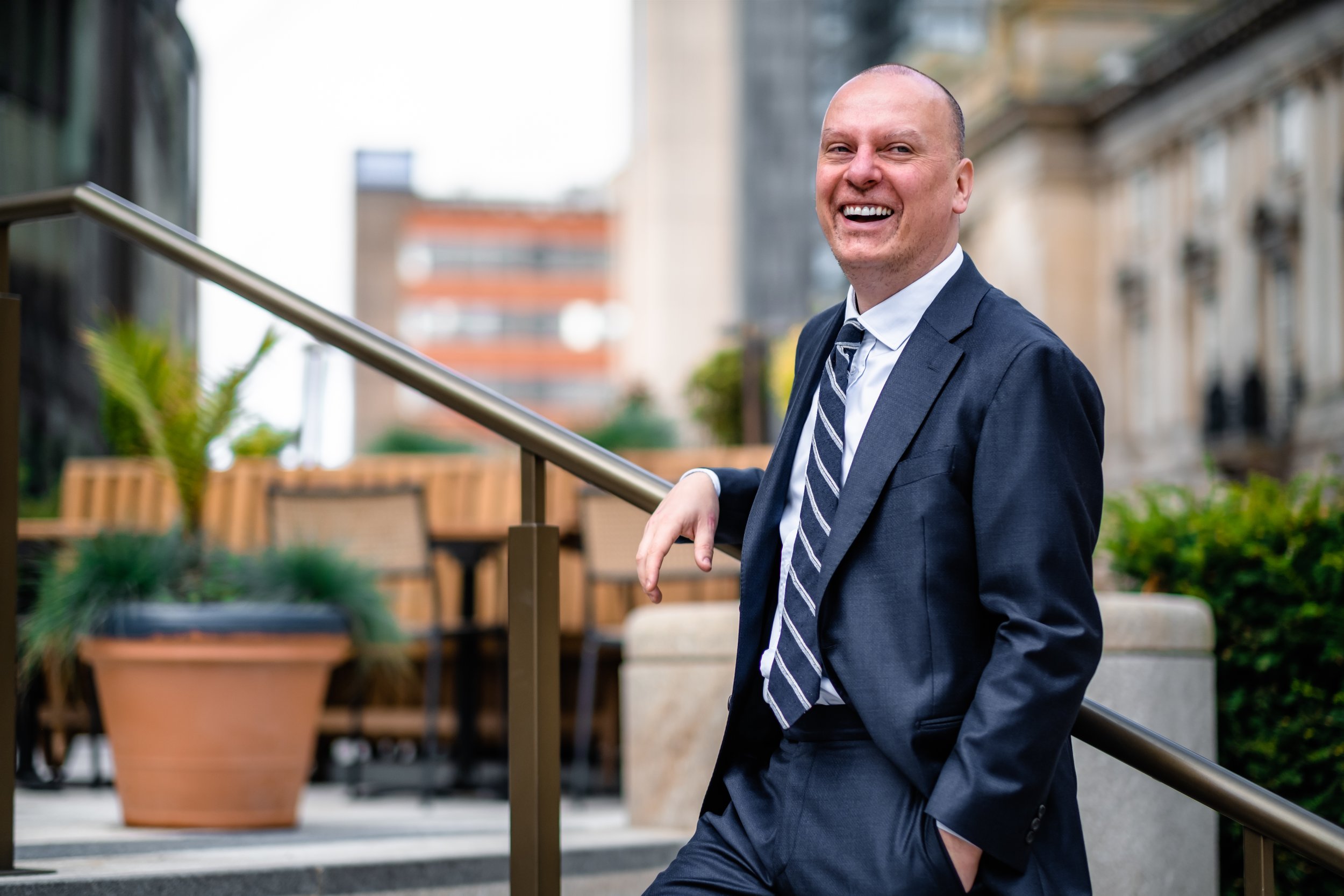 A smiling man in a business suit standing outdoors on a city street, leaning against a railing.