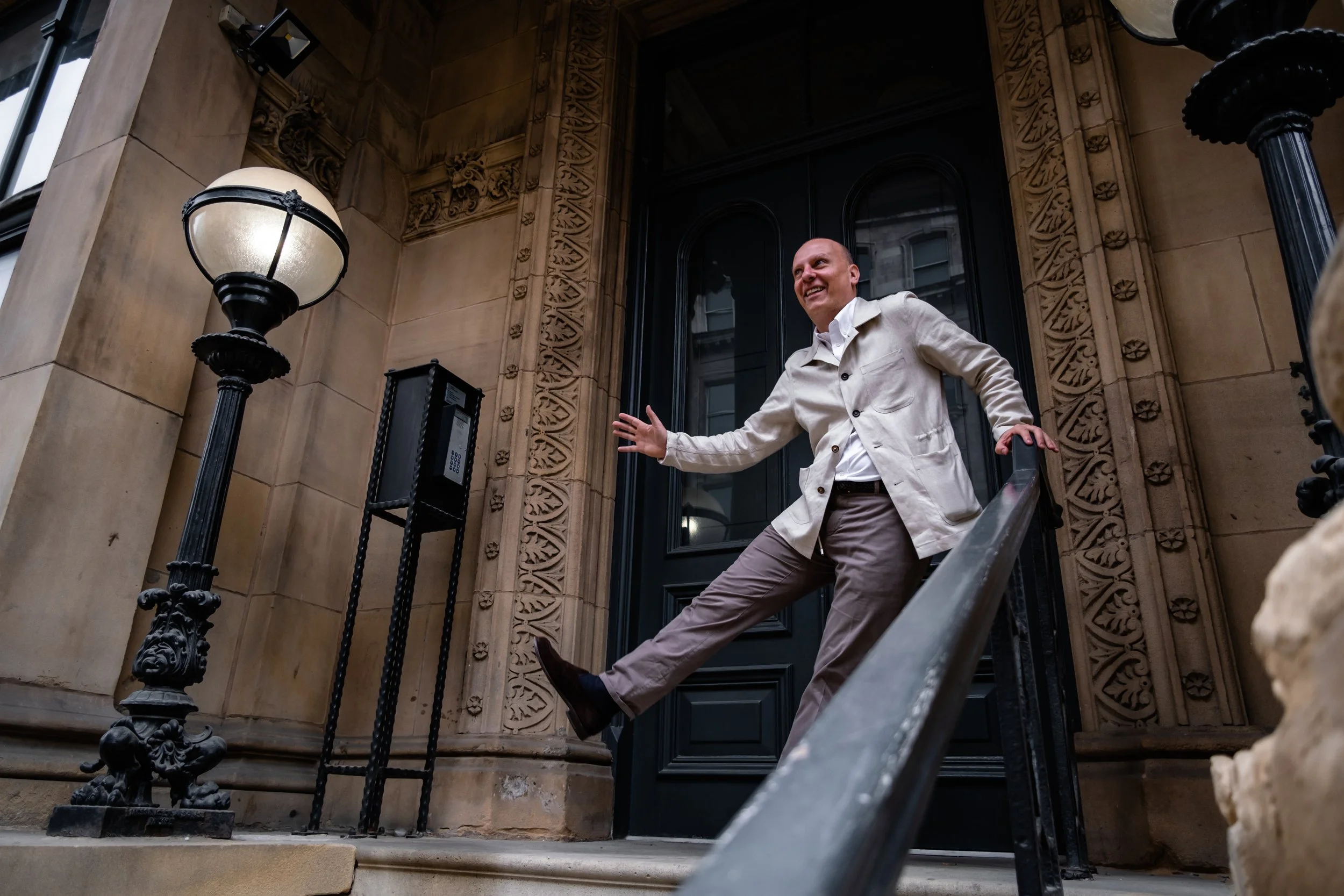 A man in a beige jacket and gray pants walking down stairs outside a building, smiling and gesturing with one hand.