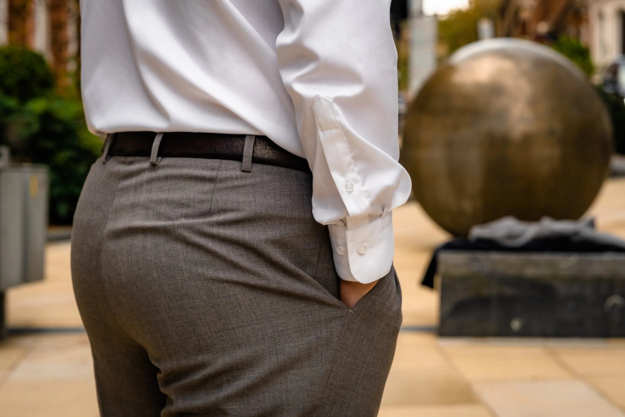 Close-up of a person wearing gray dress pants, a white dress shirt with the cuff unbuttoned, and a dark belt, standing outside near a spherical bronze sculpture.