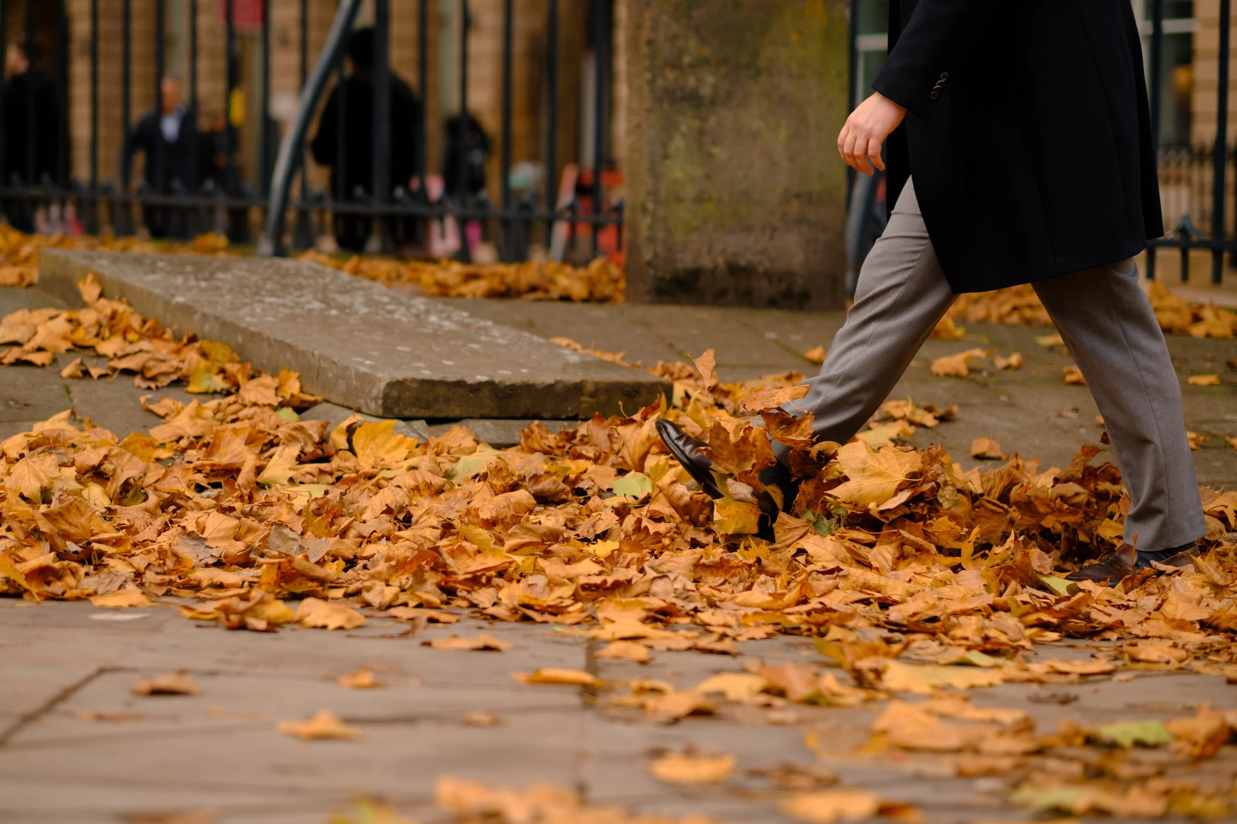 Person in business attire walking through fallen autumn leaves on a city sidewalk.