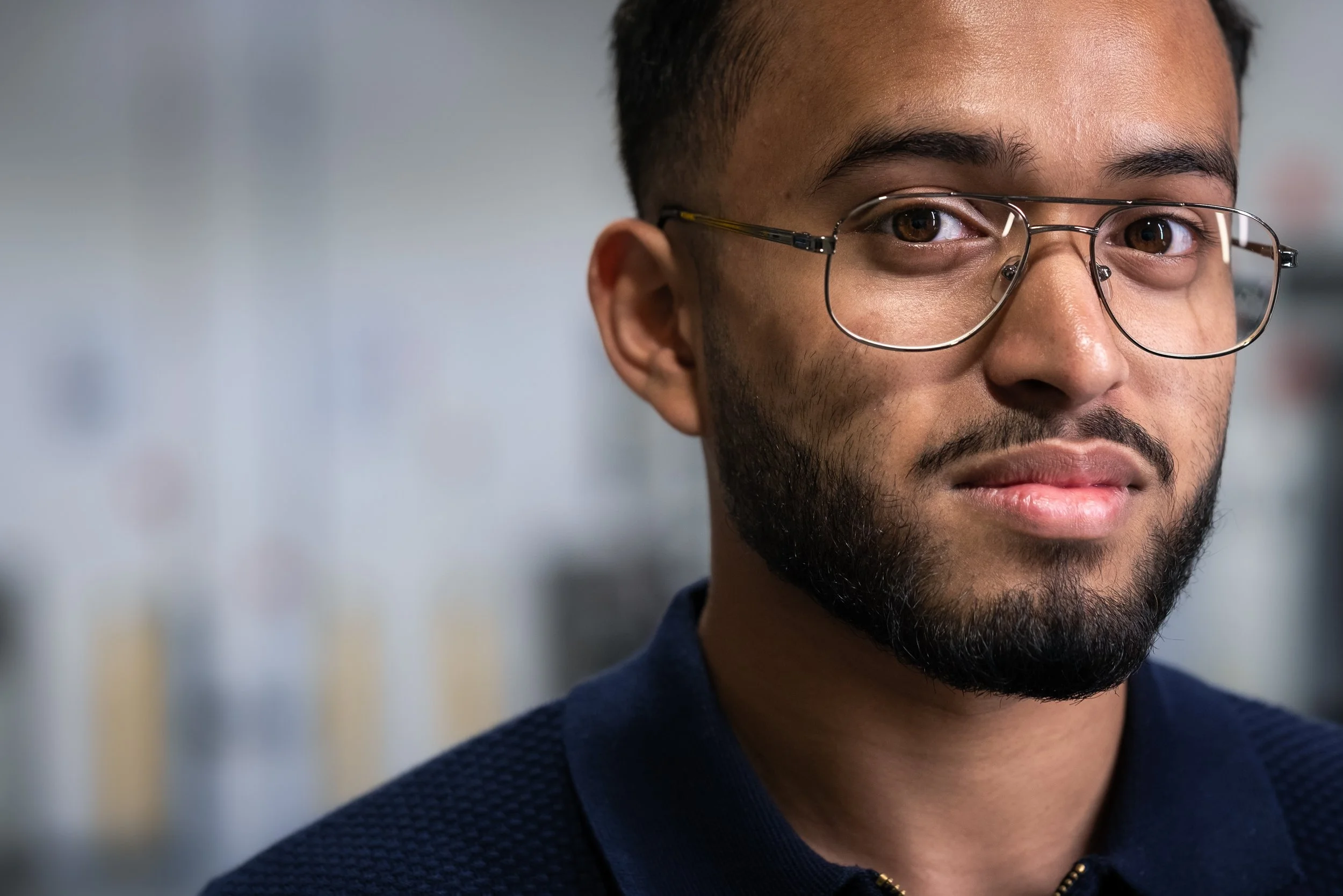 Close-up of a young man with dark hair, glasses, a beard, and wearing a navy blue sweater, standing indoors with a blurred background.