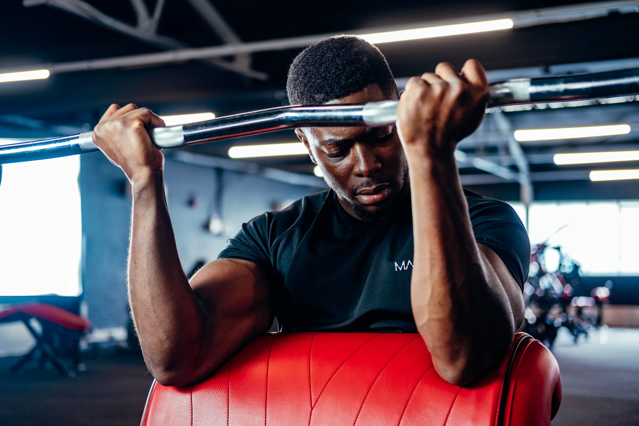A muscular man lifting a barbell in a gym, resting his elbows on a red padded support, with gym equipment and large windows in the background.