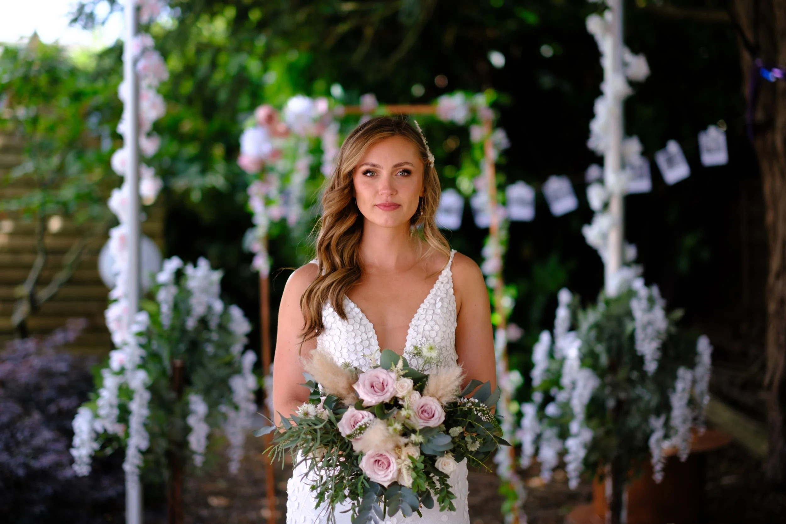 A bride holding a bouquet of pink roses and greenery at an outdoor wedding, with floral decorations and photos hanging in the background.