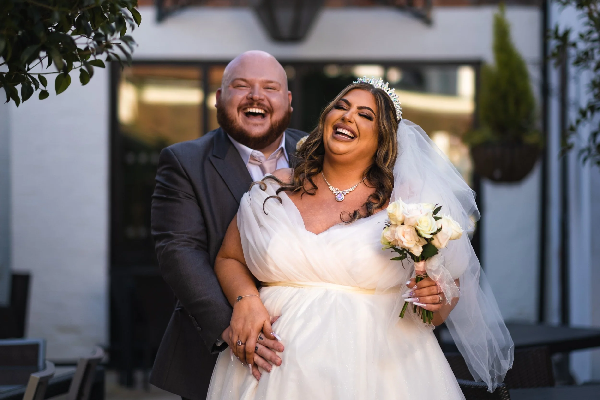 A joyful couple, a bride and groom, laughing together outdoors during their wedding celebration. The bride wears a white wedding gown, a tiara, and a necklace, holding a bouquet of roses. The groom is dressed in a dark suit and white shirt. They are holding hands and seem very happy.