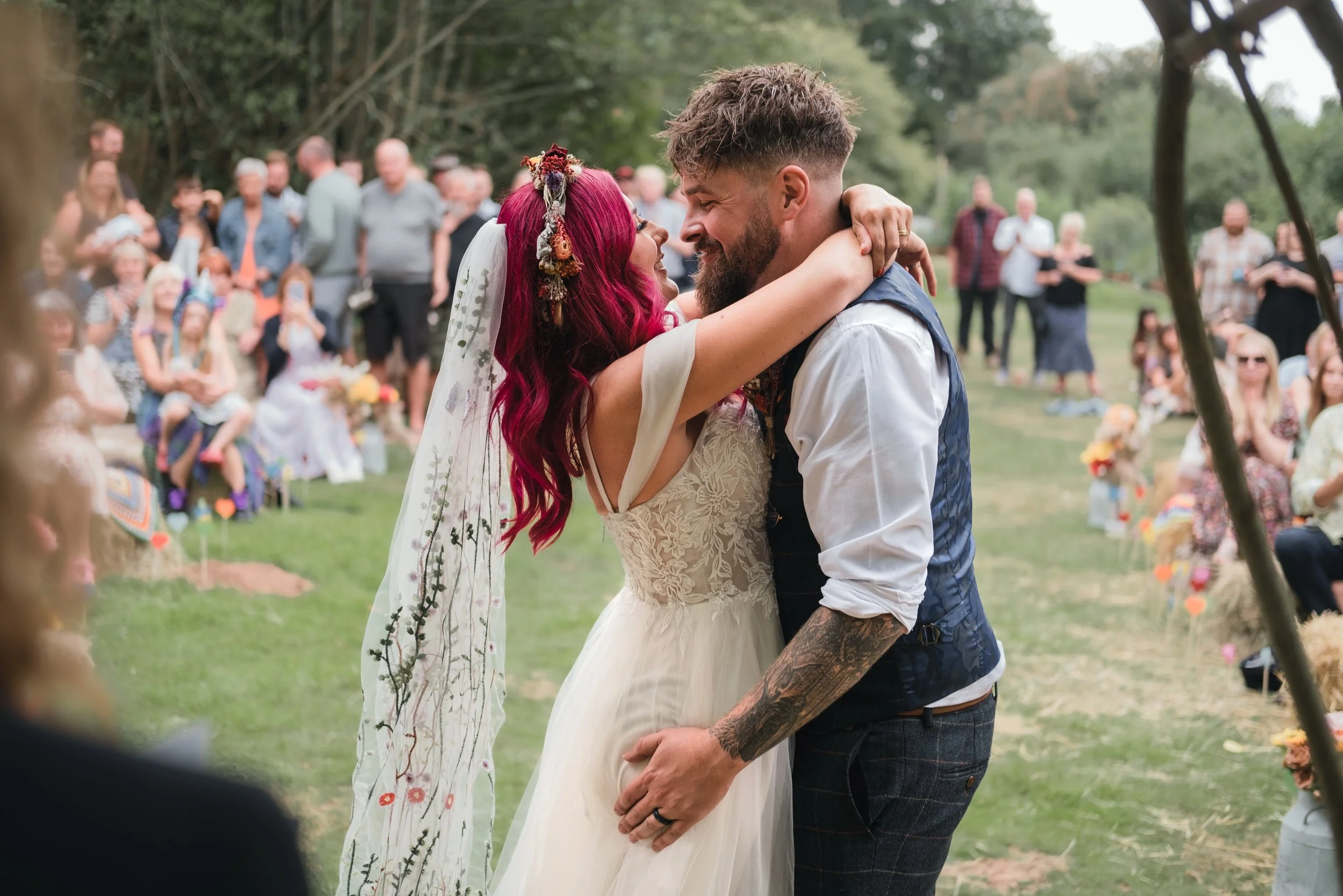 A couple shares a joyful embrace during their outdoor wedding ceremony, surrounded by friends and family seated on the grass.