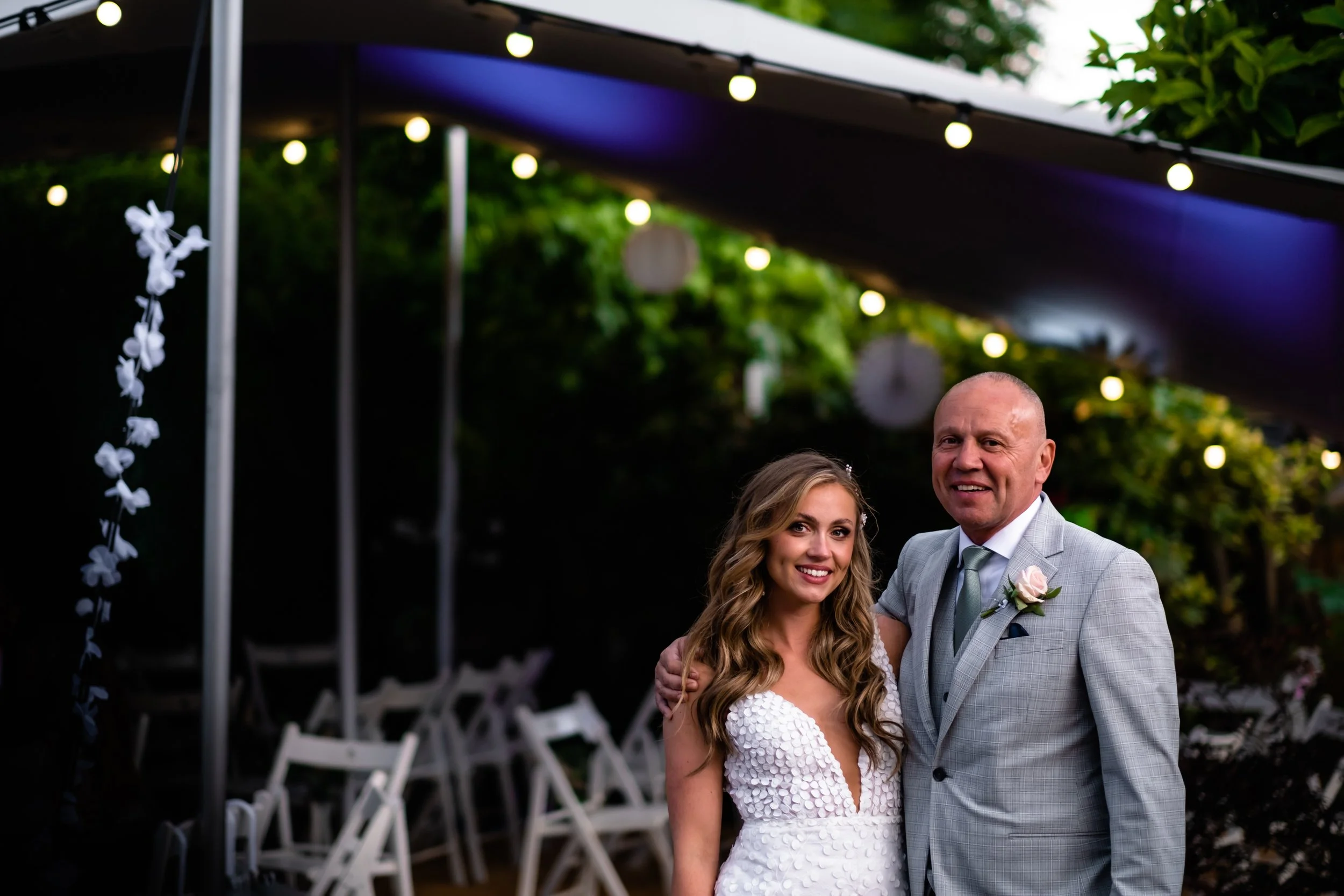 A smiling bride and groom standing outdoors under string lights and a dark canopy, with white chairs in the background, during their wedding celebration.