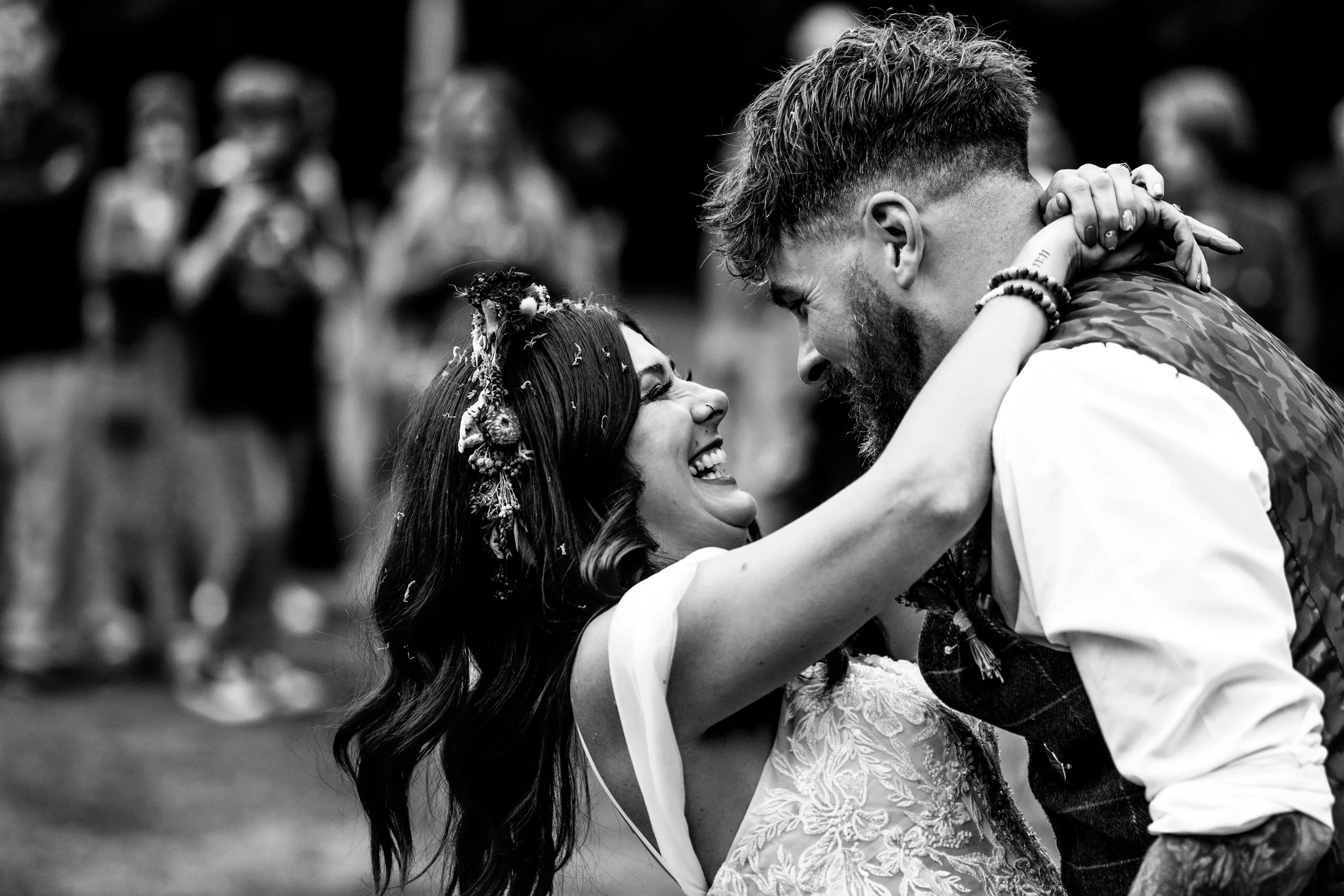 A couple shares a kiss and dance at a wedding, with the woman's arms around the man's neck, surrounded by blurred onlookers in the background.