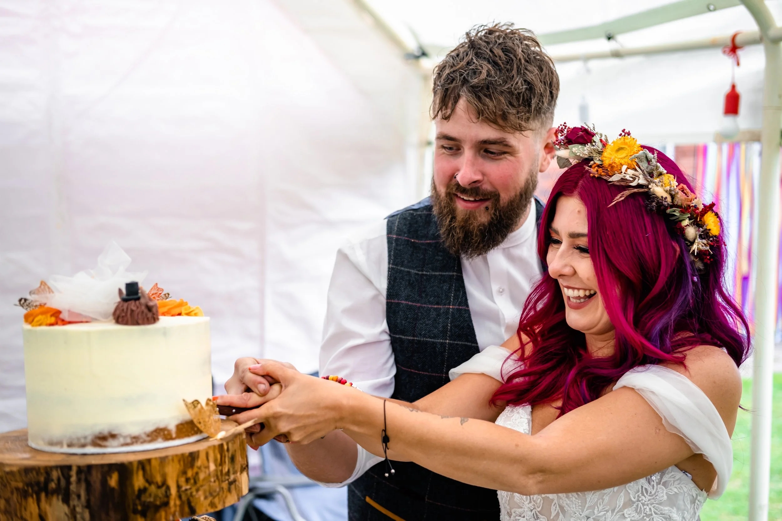 A couple cutting a wedding cake together, smiling, at a celebration. The woman has long, vibrant pink and purple hair and wears a floral crown, while the man has short, curly hair and a beard. The cake is on a wooden stand, decorated with flowers and small figures.