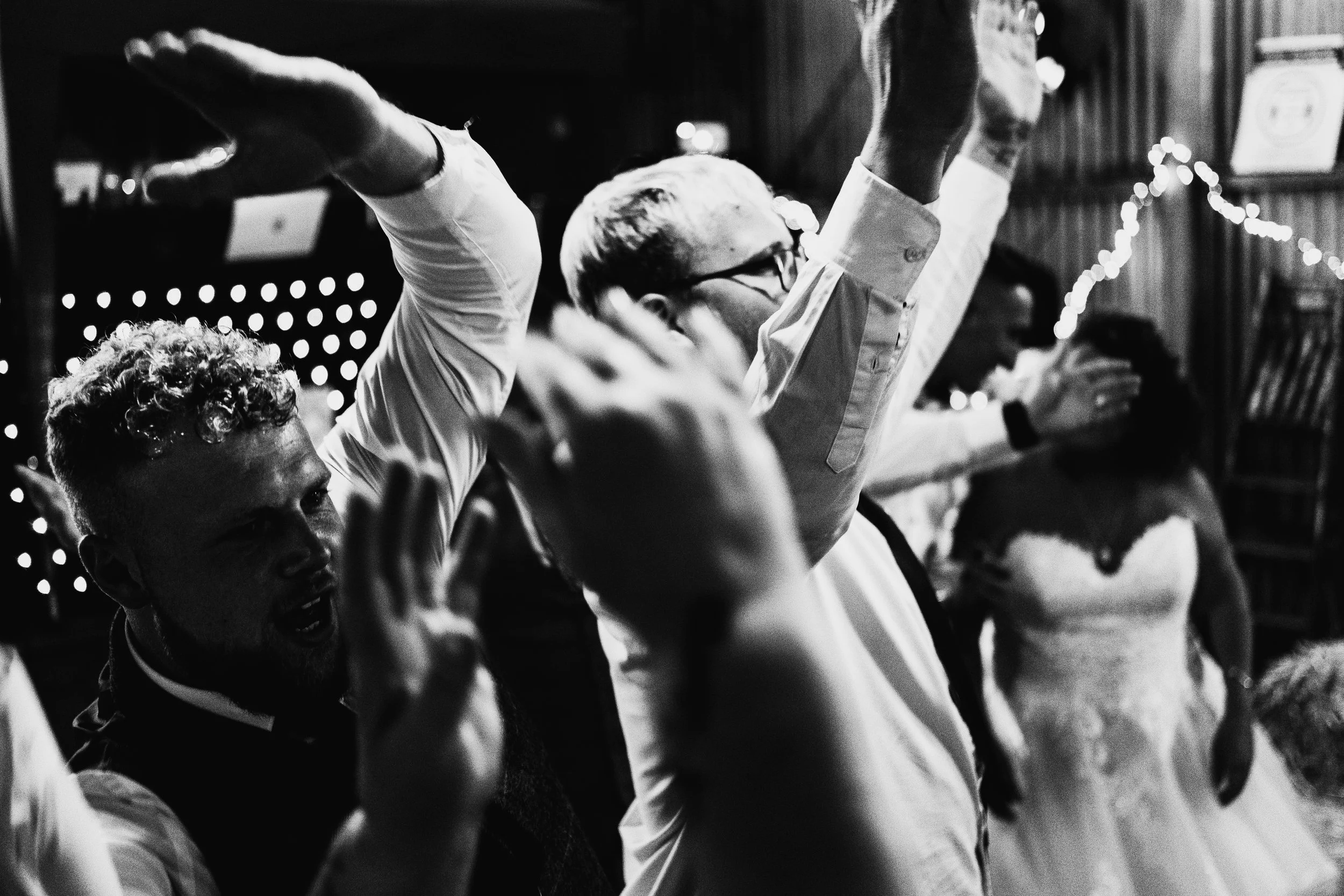 Black and white photo of people dancing at a celebration, including a man in glasses raising his arms and a woman in a wedding dress.