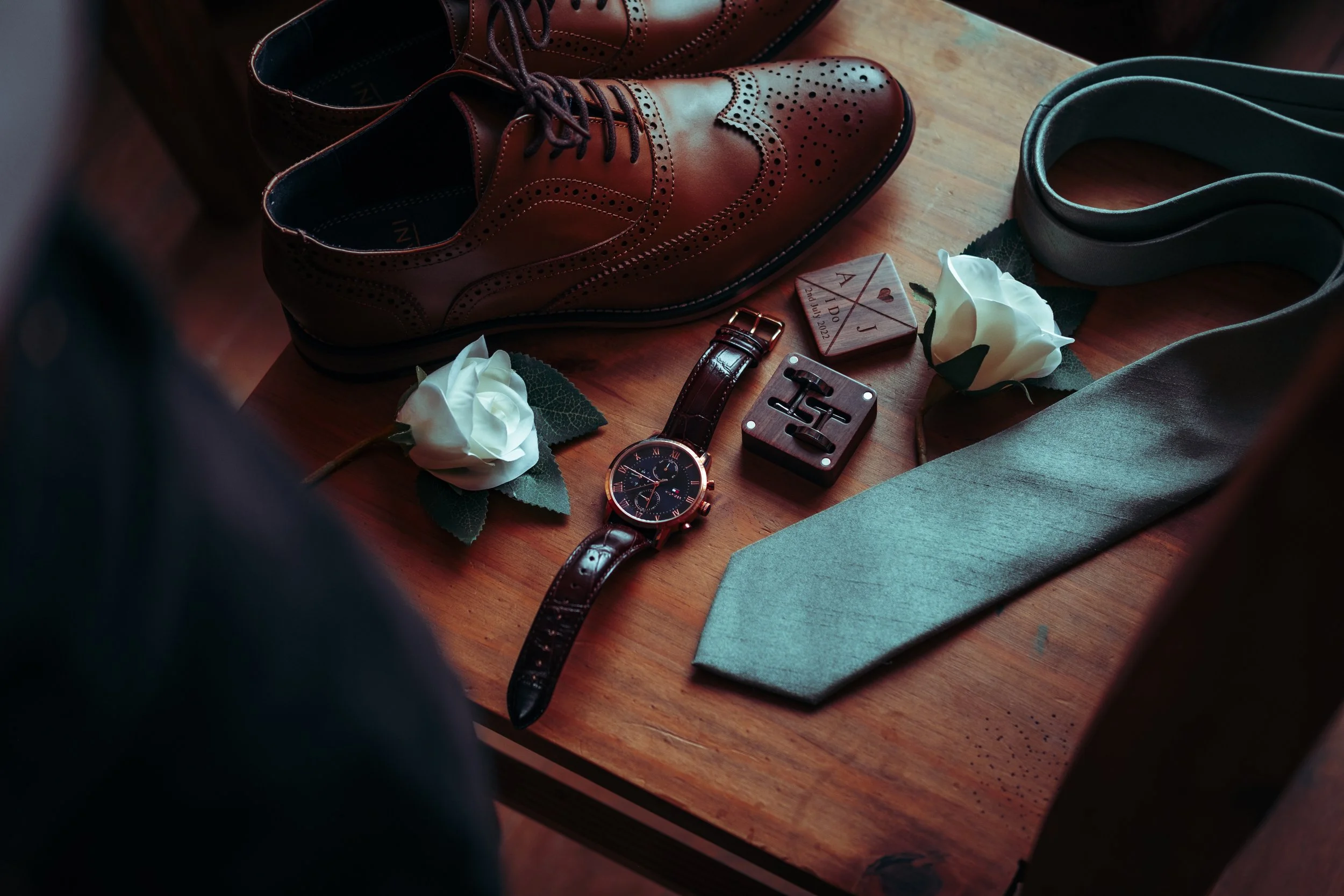 A pair of brown leather dress shoes, a gray necktie, a wristwatch with a leather strap, two white roses, a deck of playing cards, a wooden box, and a wooden trinket are arranged on a wooden surface.