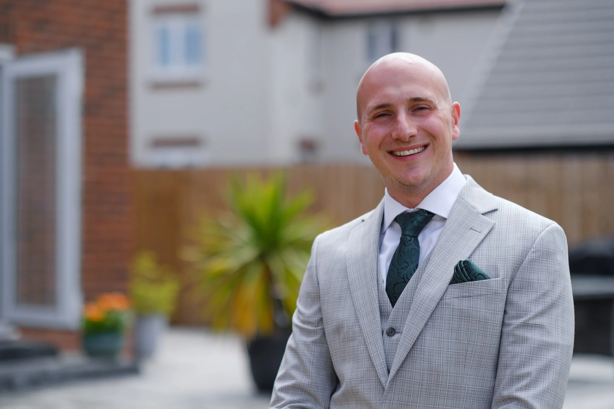 A smiling man in a light gray suit with a white shirt and dark green tie standing outdoors in front of a house with blurred background.