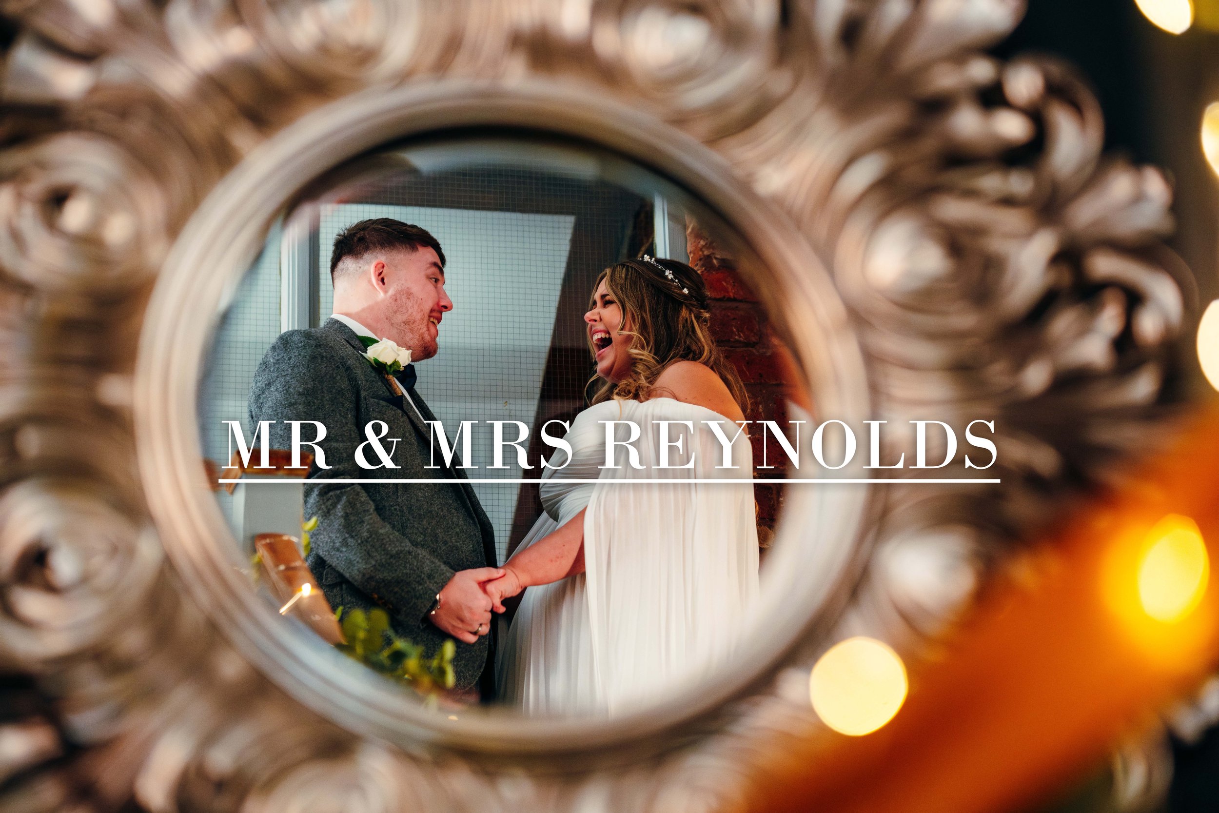 A bride and groom holding hands and smiling at each other during their wedding ceremony, seen through a decorative mirror with the text 'MR & MRS REYNOLDS' overlaid.