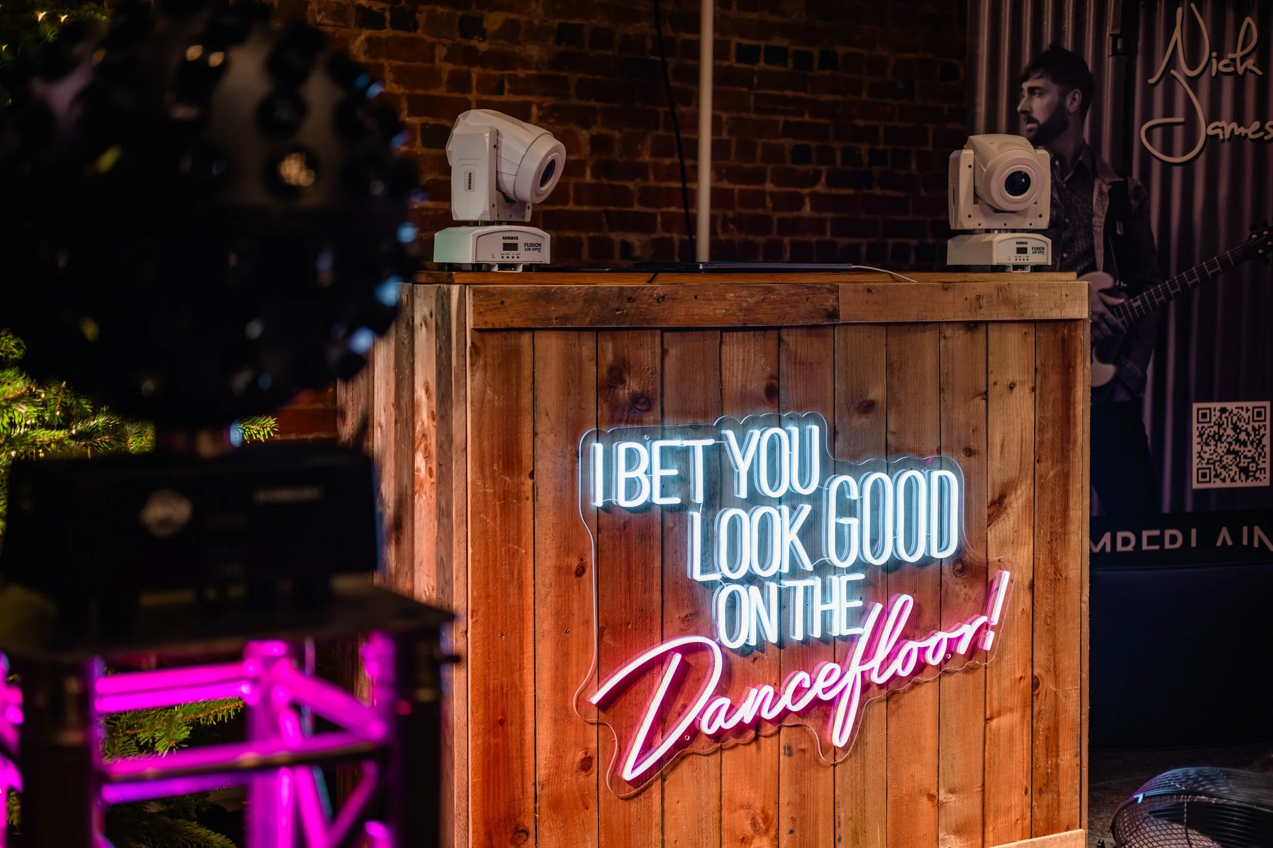 Neon sign on a wooden bar that says 'I BET YOU LOOK GOOD ON THE DANCEFLOOR' in a dimly lit setting, with a person and DJ equipment in the background.