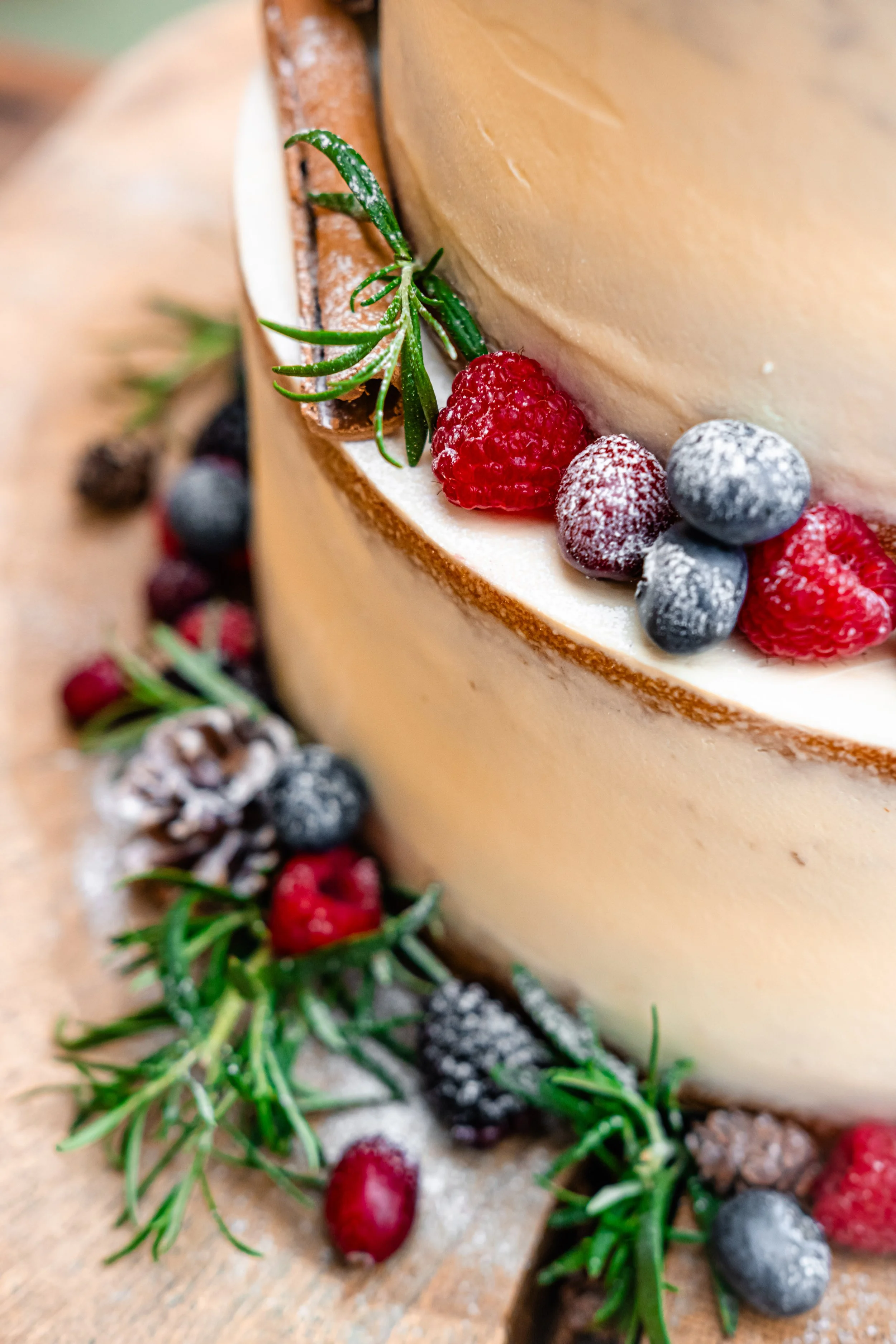 Close-up of a frosted Christmas-themed cake decorated with raspberries, blueberries, pine cones, cinnamon stick, and sprigs of rosemary on a wooden surface.