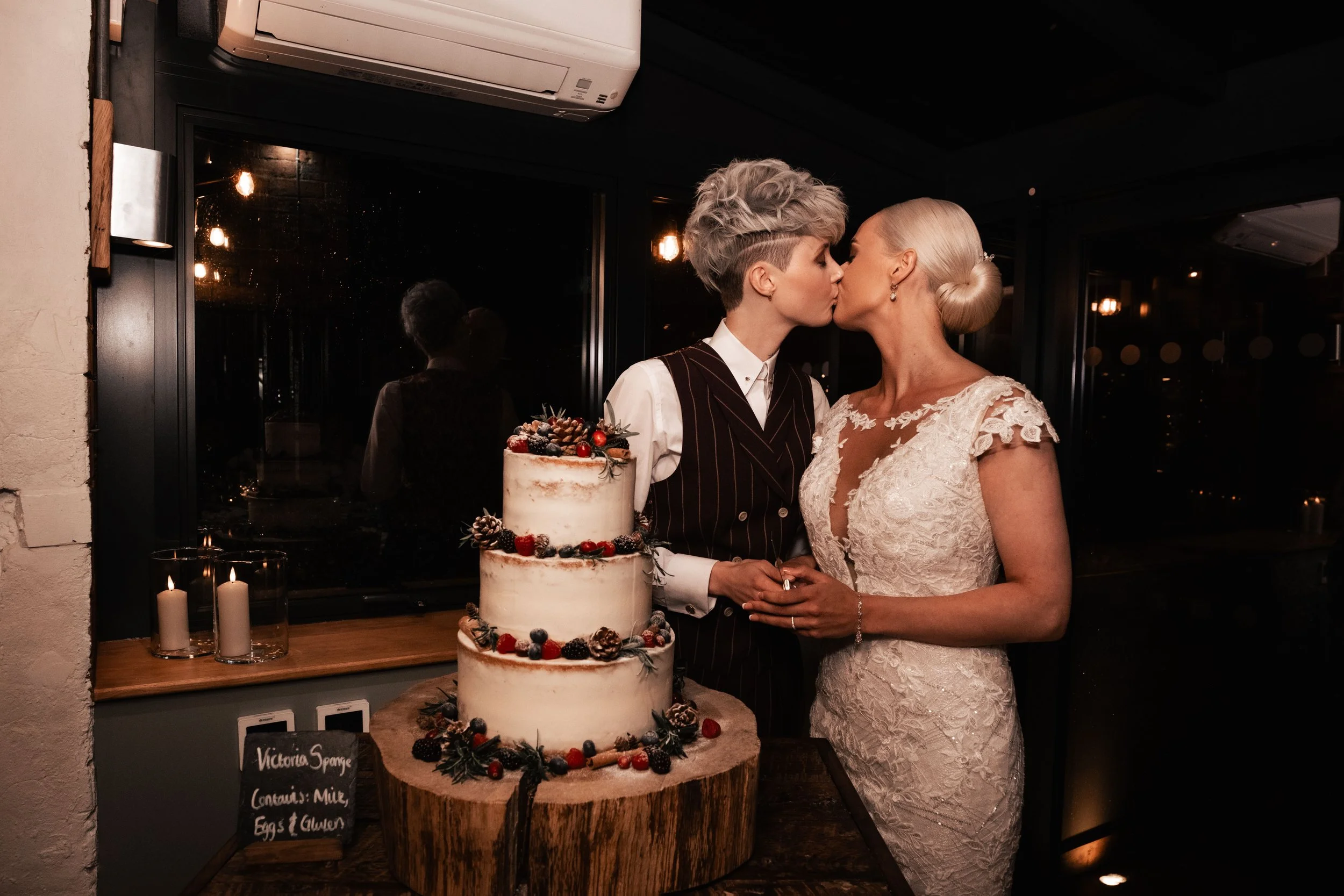 Two women, dressed in wedding attire, share a kiss beside a three-tiered wedding cake decorated with berries and pinecones, in a dimly lit indoor setting.